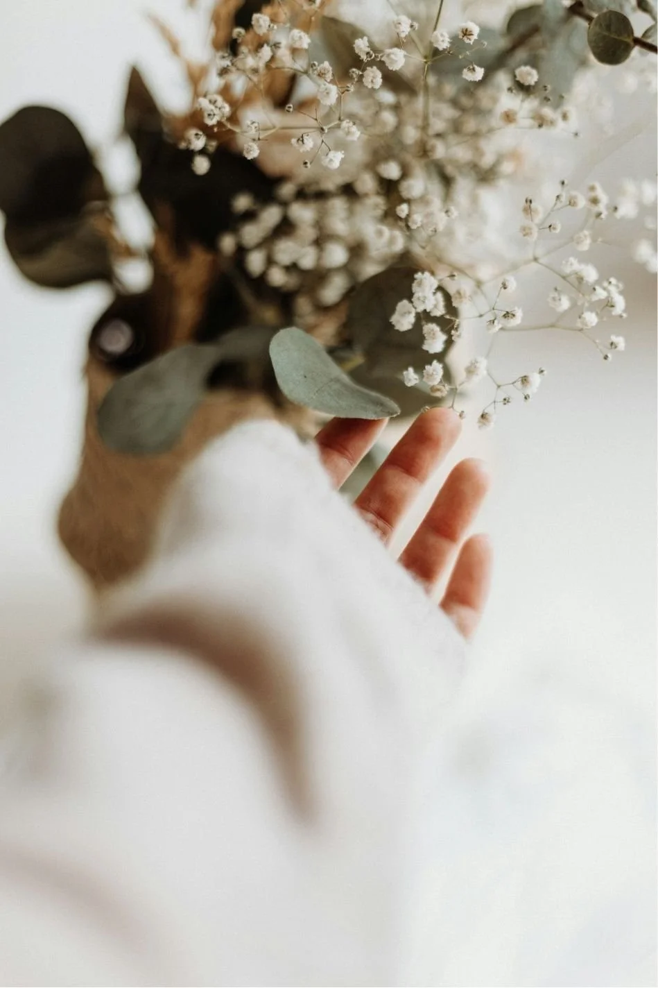 A hand reaching towards a bouquet of small white flowers and eucalyptus leaves, part of a floral arrangement.