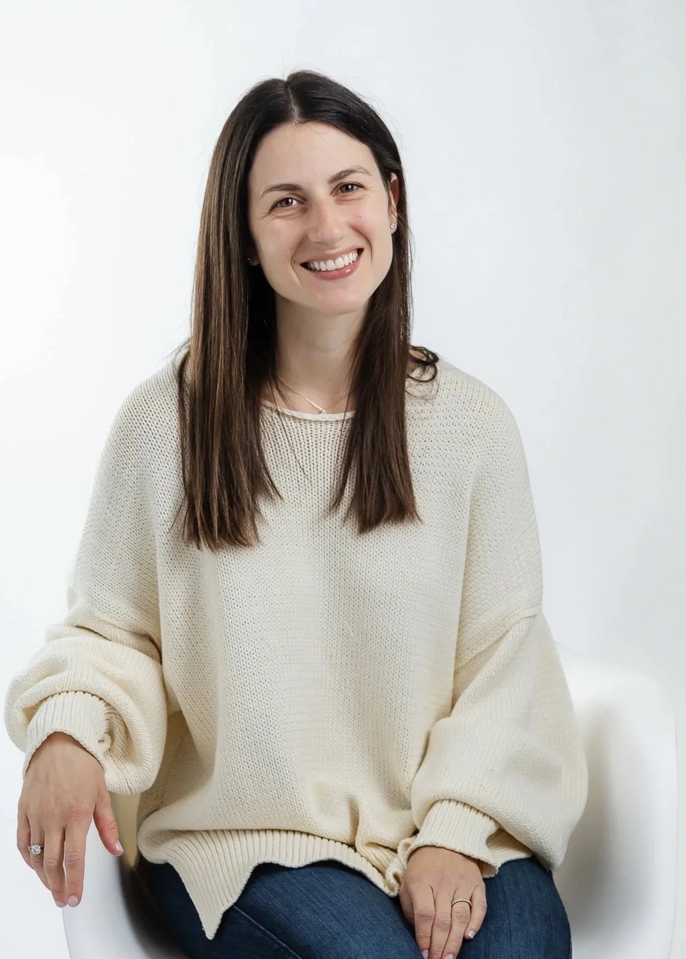 A woman with long brown hair sitting on a white chair, smiling, against a plain white background, wearing a cream sweater and dark jeans.