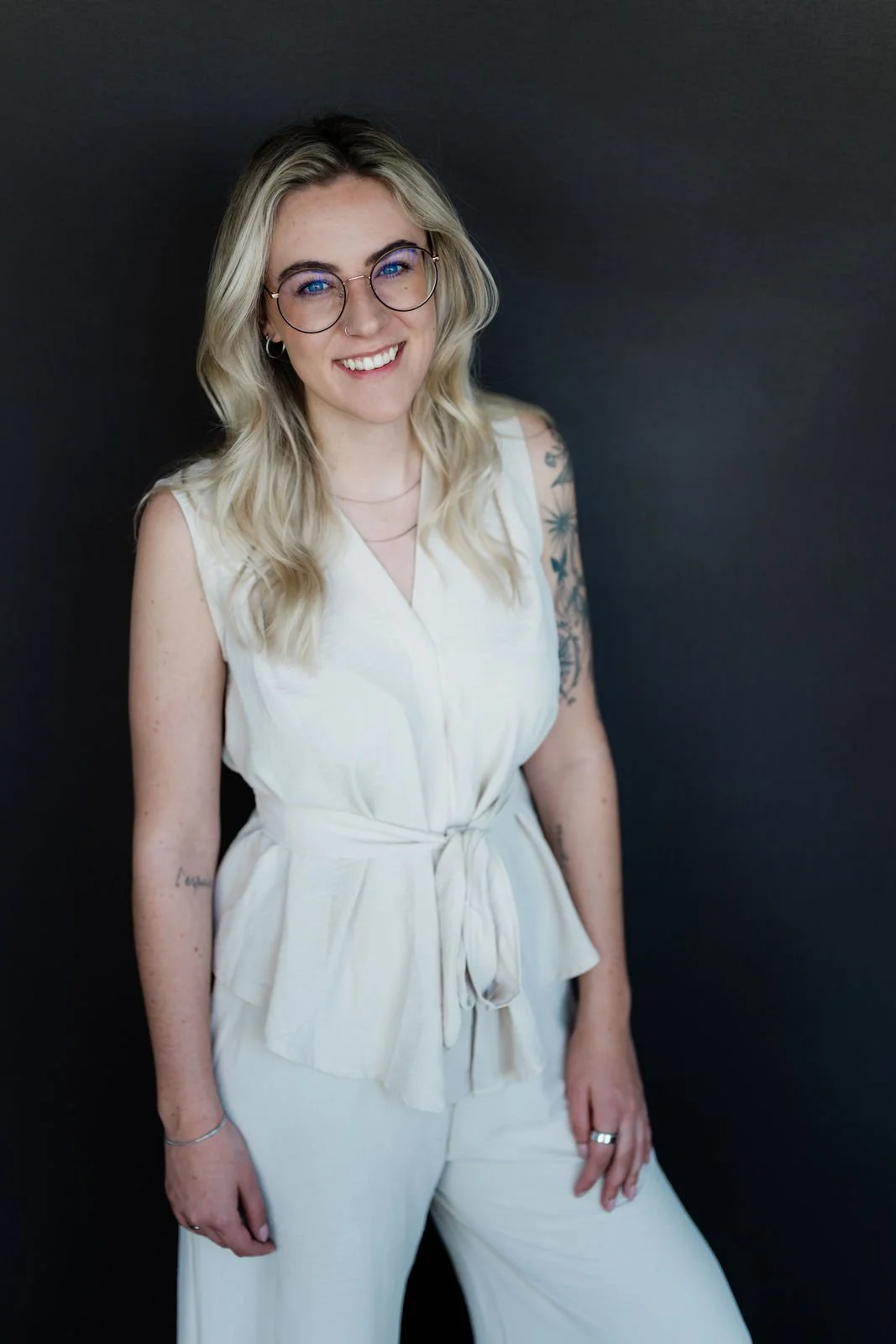 Junge Frau mit Brille und Tattoos, trägt weiße Kleidung, lächelt vor dunklem Hintergrund.