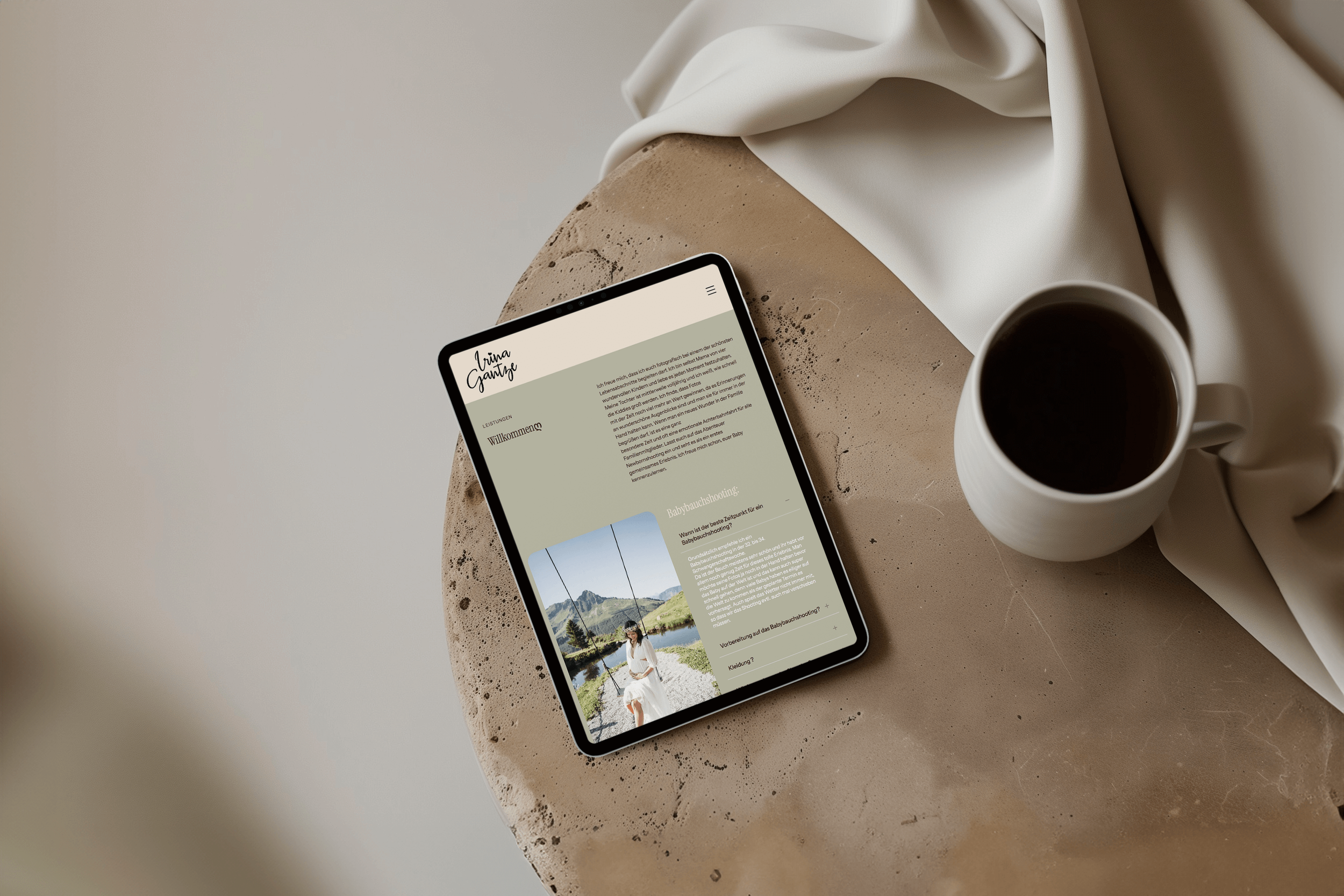 Tablet auf einem runden, beigefarbenen Tisch mit weißer Kaffeetasse und weißem seidigem Tuch im Hintergrund, zeigt eine Website mit Text und Foto einer Frau vor Berglandschaft.