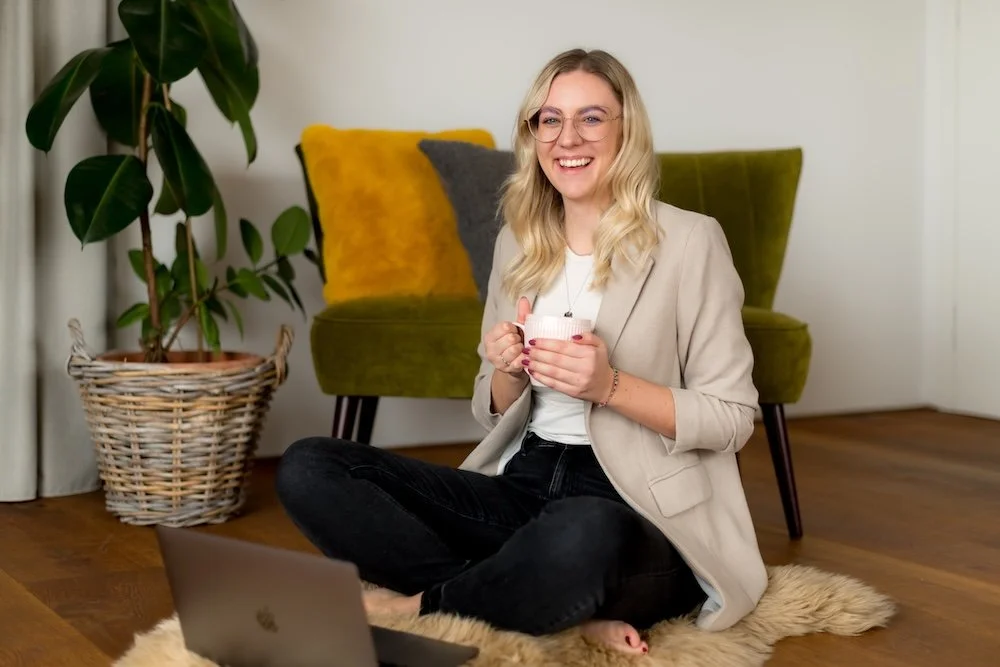 Eine lachende Frau sitzend auf einem Teppich, die einen Becher hält, im Hintergrund ein grüner Sessel mit Kissen, neben einer Zimmerpflanze in einem Korb, vor einem offenen Laptop.