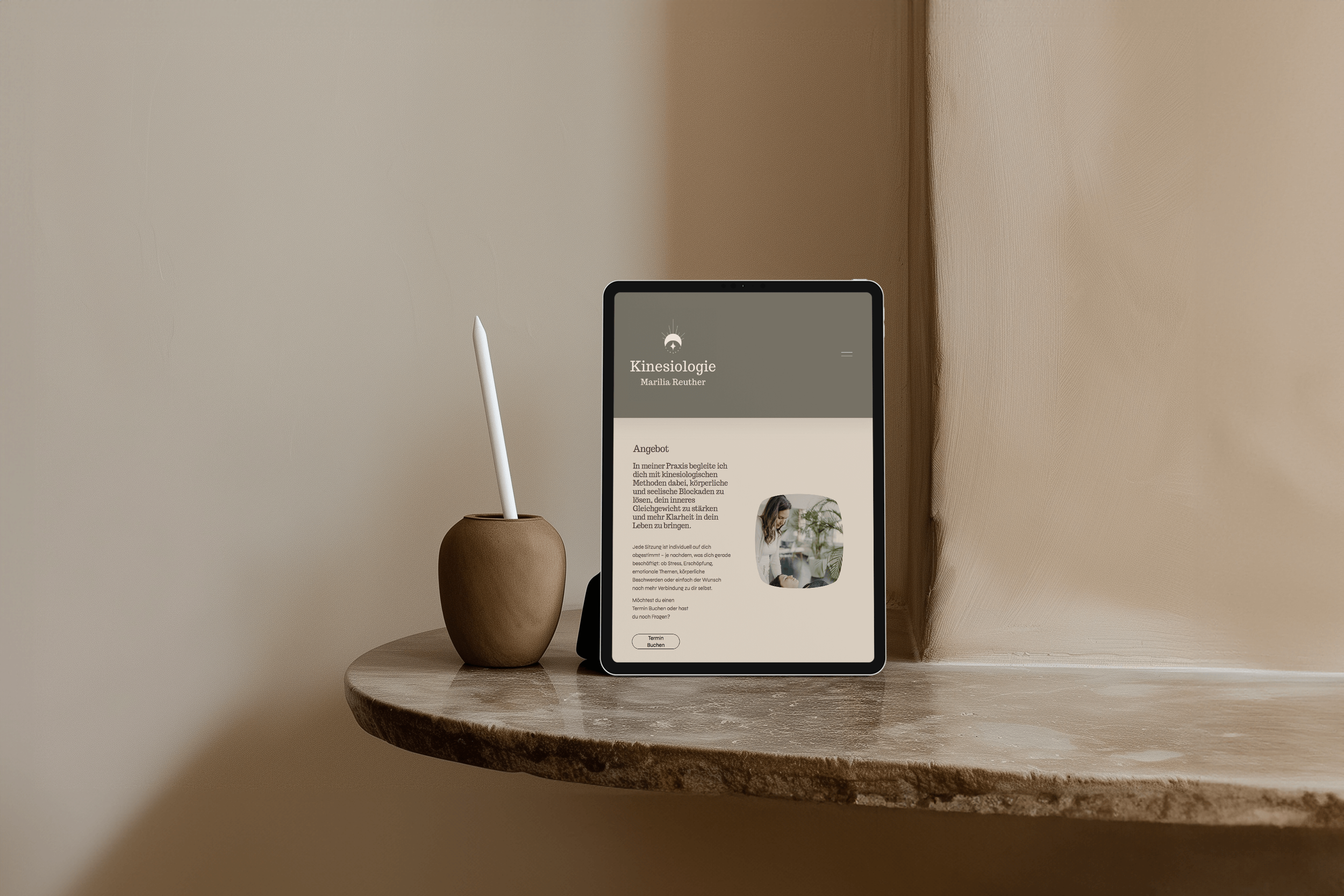 Ein Tablet zeigt eine Website für Kinesiologie, neben einem braunen Keramikvasenhalter mit einem Stift, auf einem runden Holztisch, an einer Wand in einem neutralen Raum.