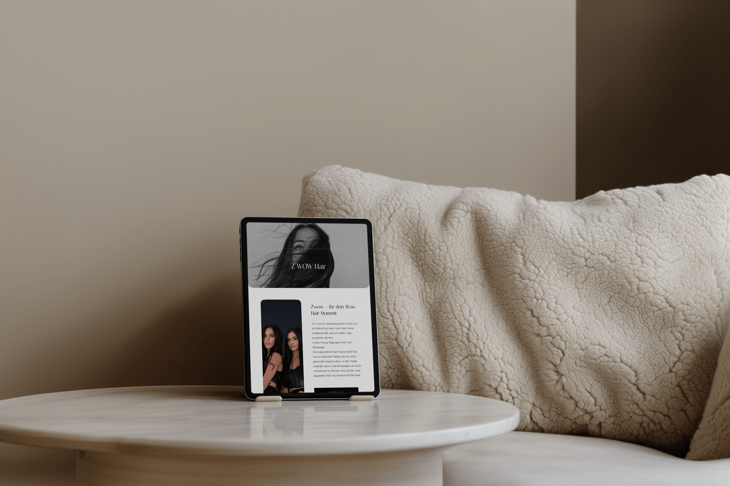Ein Tablet auf einem weißen Tisch zeigt eine Website namens 'Z'WOW Hair' mit Bildern von Frauen mit gepflegtem Haar und Text. Im Hintergrund ein beige, kuschelige Decke auf einem Sofa.