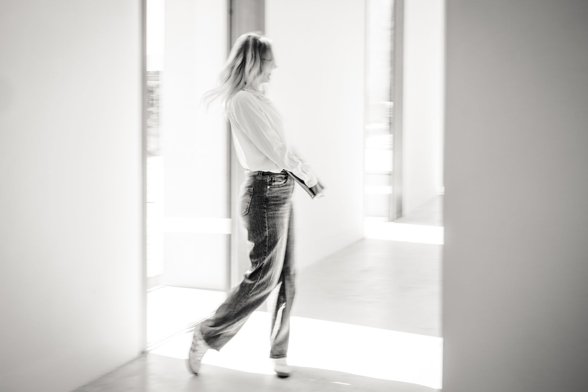 Schwarz-weiß Foto einer Frau in einem modernen, hellen Raum. Sie trägt Jeans und eine lockere Bluse, hält ein Buch in der Hand und läuft Richtung Fenster. Die Szenerie wirkt ruhig und stilvoll.