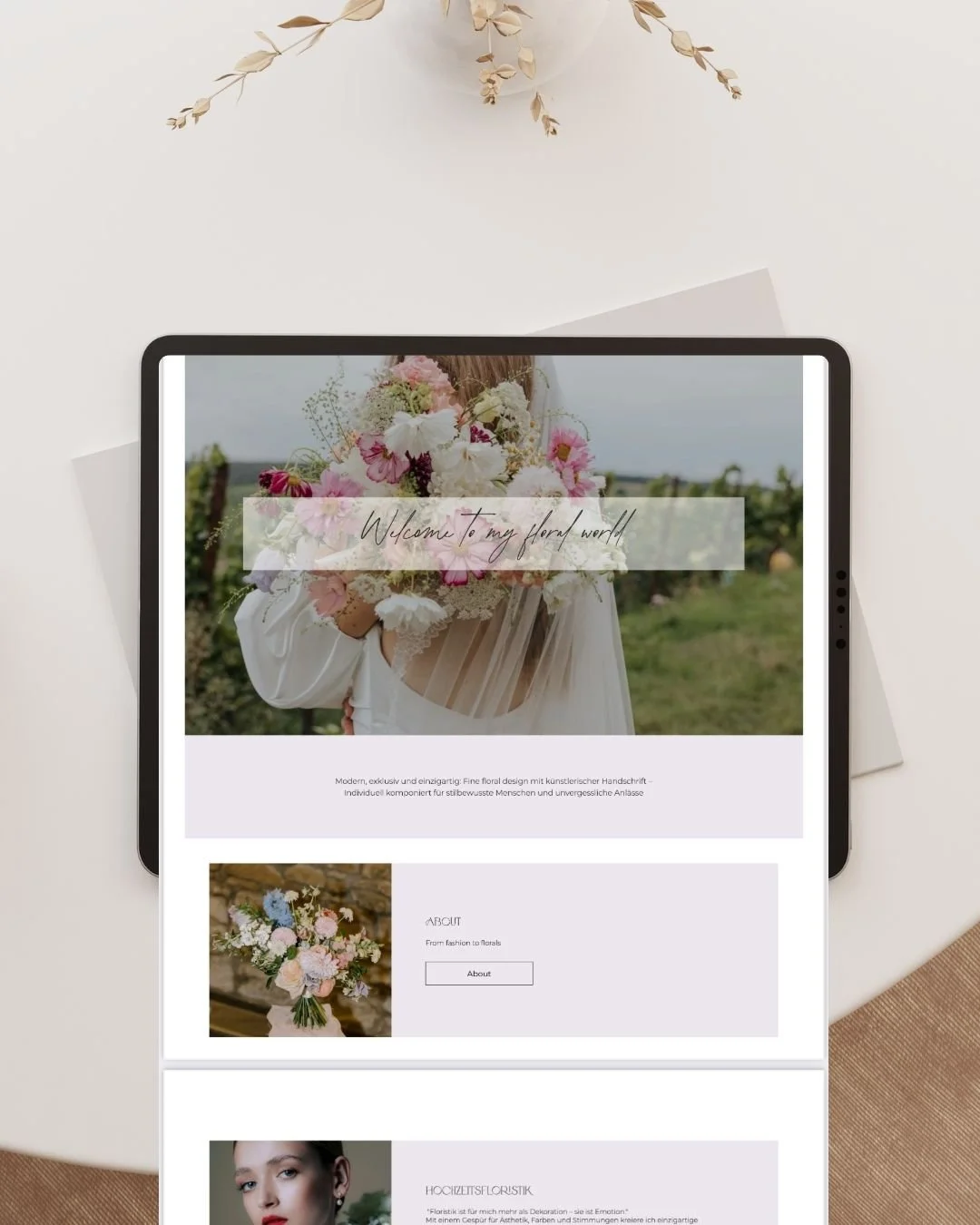 Handgehaltenes Tablet mit Florale Webseite, im Hintergrund eine weiße Tafel mit getrockneten Zweigen, auf dem weißen Tisch liegen Papiere.