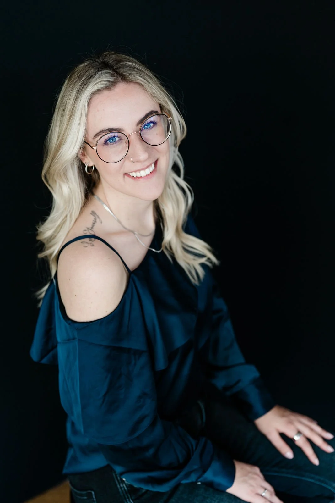 Lächelnde junge Frau mit blonden, gewellten Haaren und Brille, sitzend gegen einen schwarzen Hintergrund, trägt eine dunkle Outfits mit Shoulder-Details.