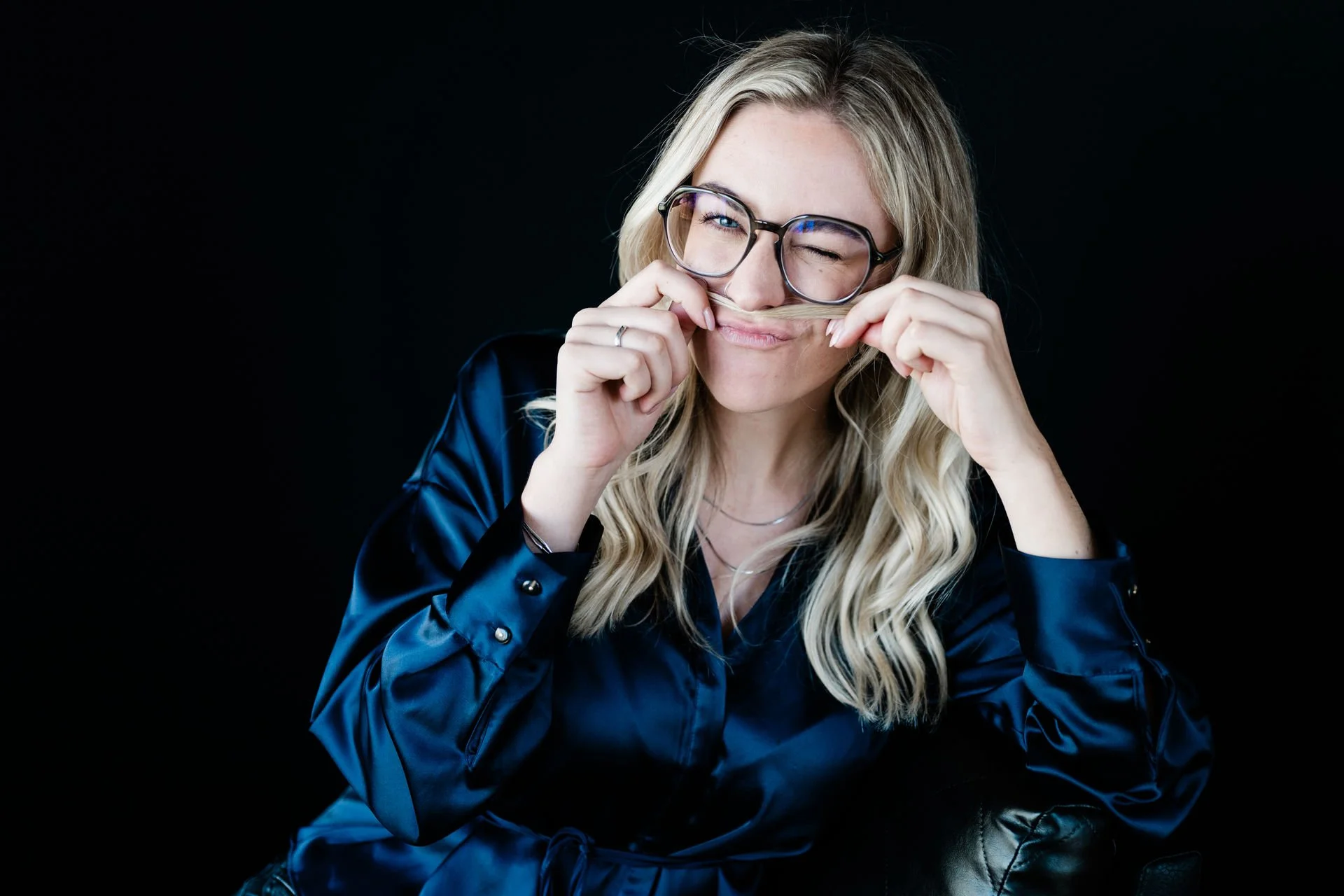 Junge Frau mit Brille spielt mit ihrem Bartstoppel und macht ein lustiges Gesicht gegen schwarzen Hintergrund.