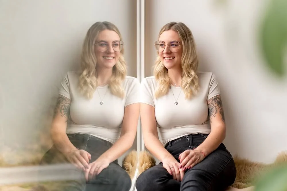 Eine junge Frau sitzt hinter einer Glasscheibe, lacht und schaut sich an, während sie sich in einem Raum mit natürlichem Licht befindet.