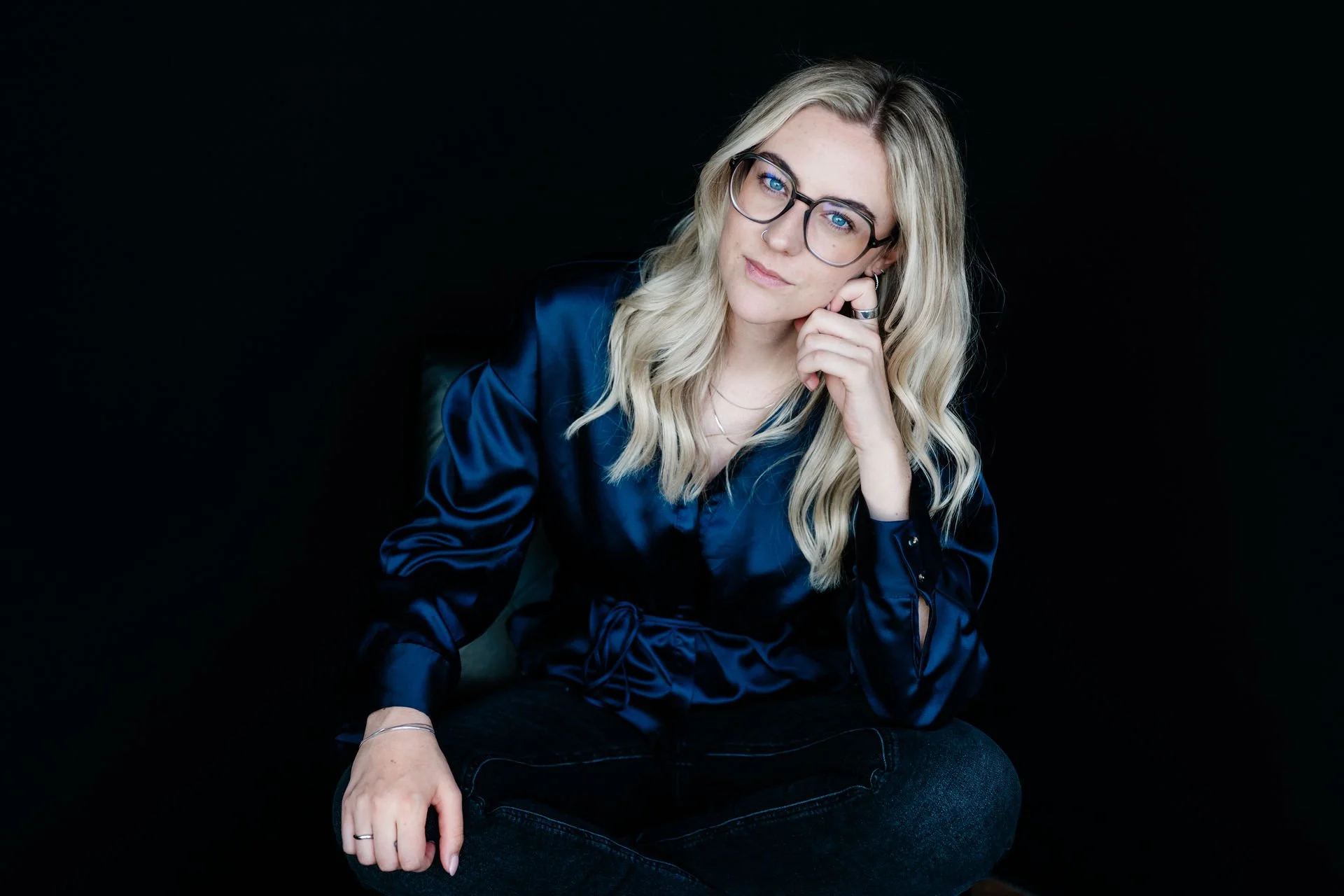 Junge Frau mit Brille und blonde, gewellte Haare, in dunklem Raum sitzend, trägt eine blaue Seidenbluse und schwarze Jeans, wirkt nachdenklich.