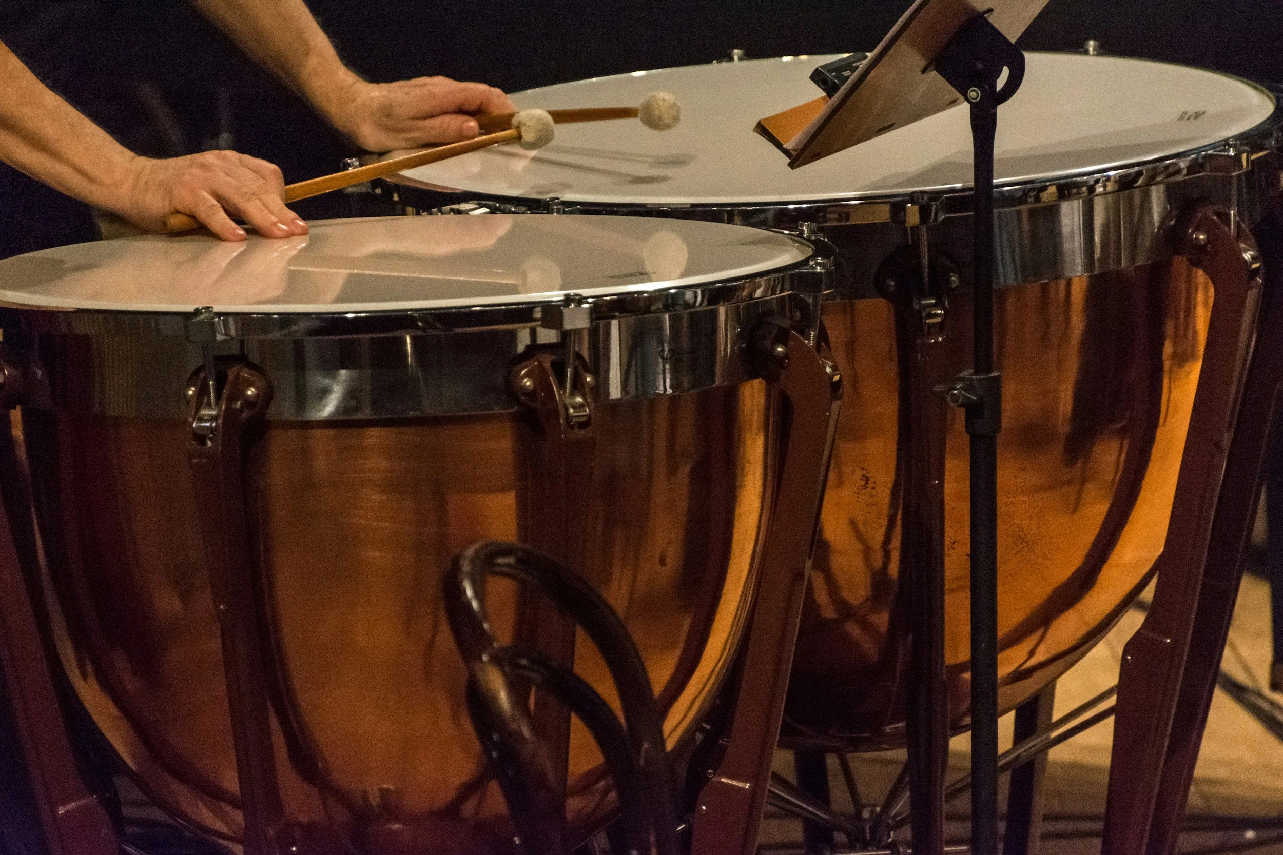 Two large timpani drums being played by a musician. The musician's hands and mallets are visible, and there's a music stand nearby.