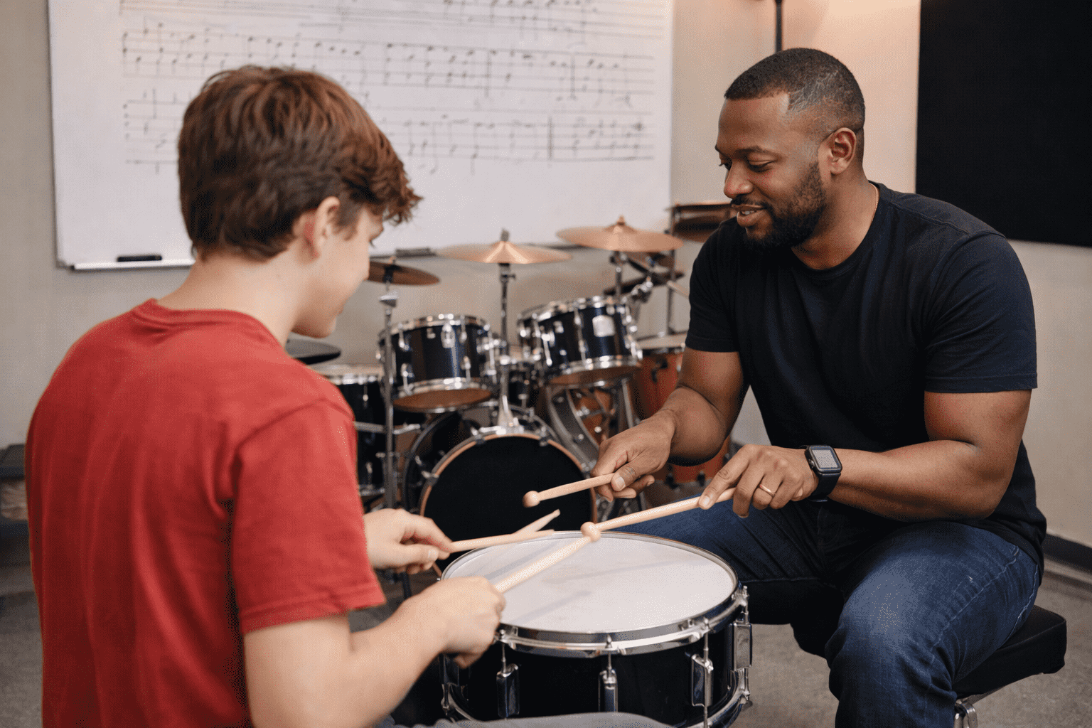 A music instructor teaching a young student how to play the drums in a music classroom with drums and sheet music on the whiteboard.
