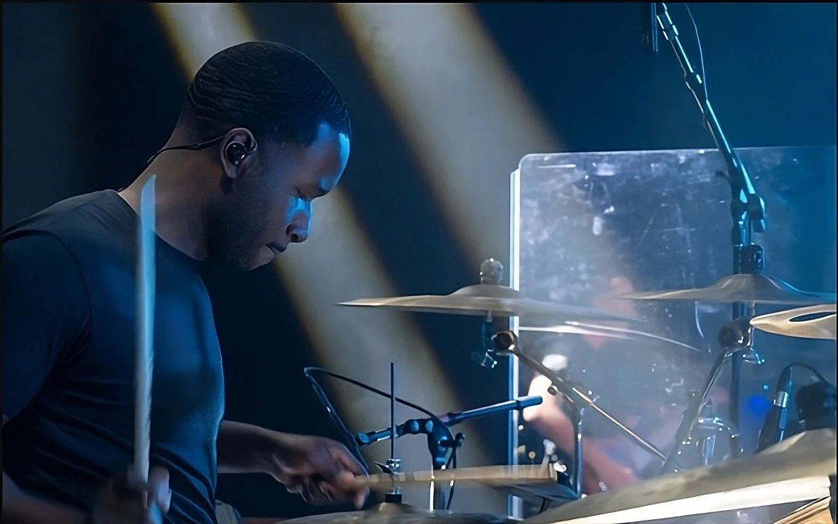 A man playing drums on stage with cymbals, bass drum, and drumsticks, with stage lighting and a transparent shield in the background.