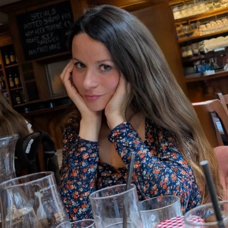 A woman with long brown hair and blue eyes sitting at a restaurant table, resting her face on her hands, wearing a floral long-sleeve shirt, with glasses and a drink in front of her, and a wooden bar in the background.