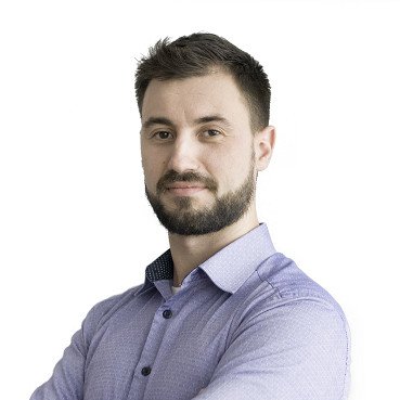 A man with dark hair and a beard wearing a light purple button-up shirt, posing with arms crossed against a white background.