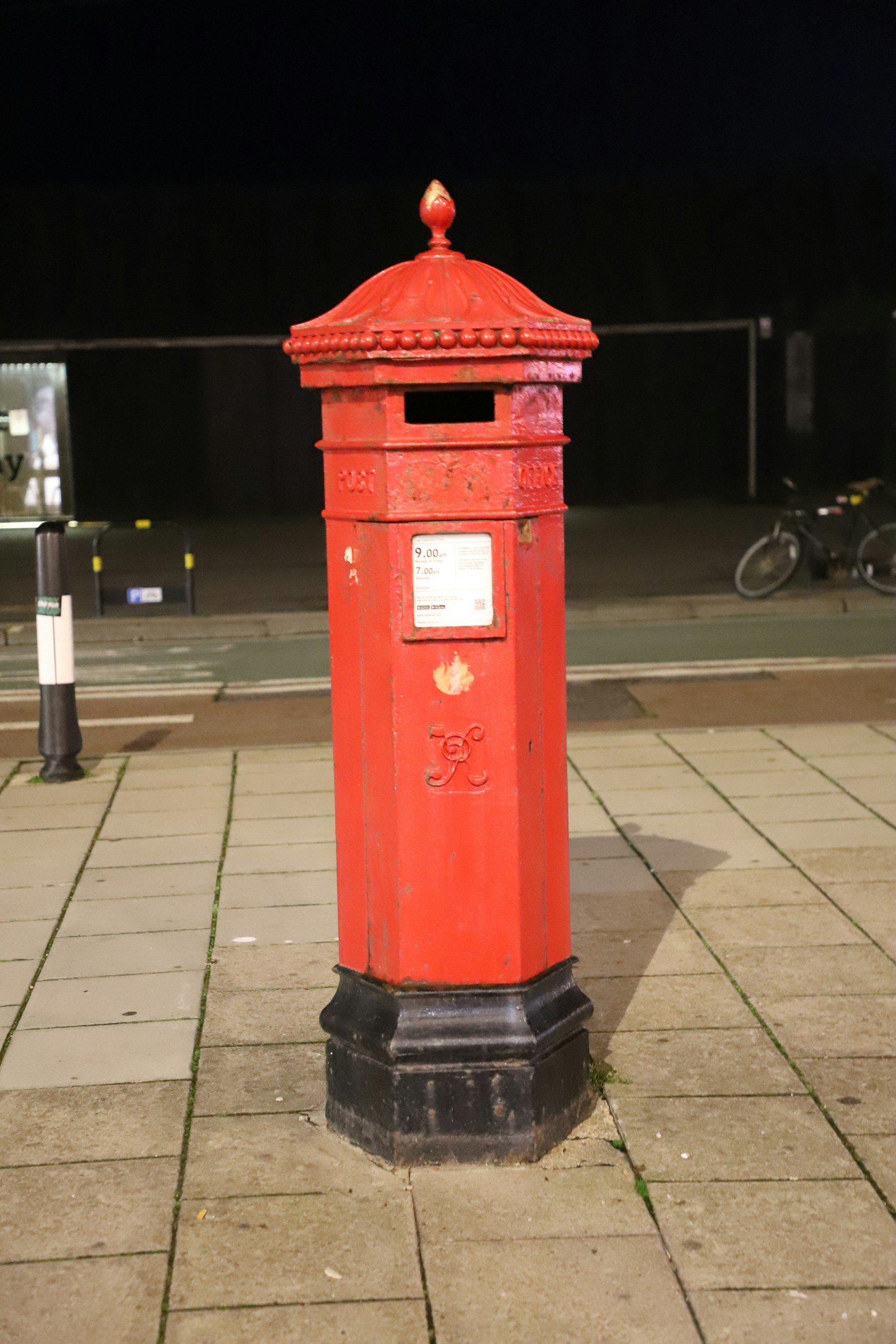 A red vintage mailbox standing on a city sidewalk at night, with a bicycle in the background and street markings visible.