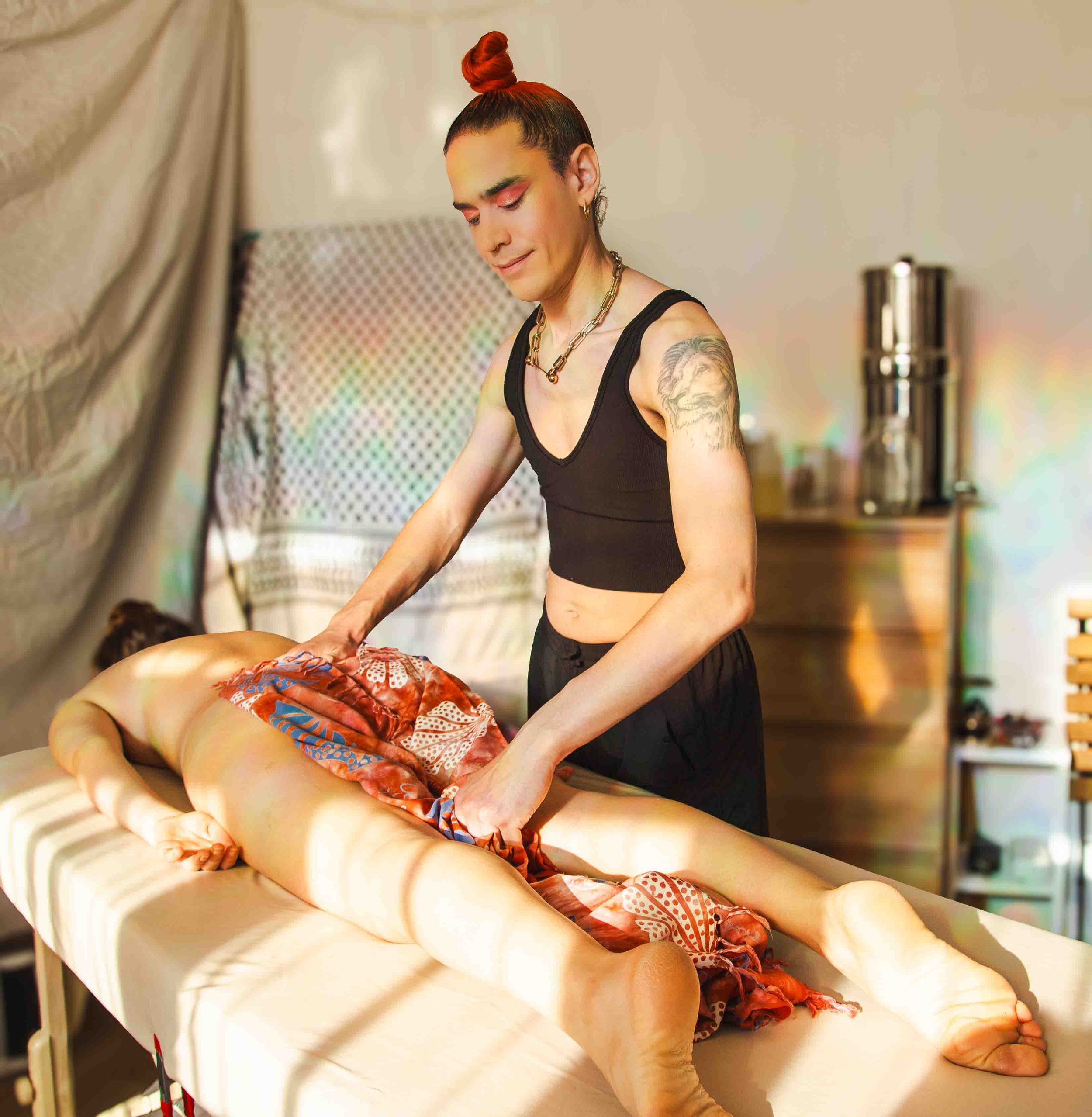 Person receiving a lomi massage on a massage table, lying face down. The masseuse, wearing a black top, is applying pressure to the person's back. The room has warm lighting, a wooden dresser in the background, and a keffiyeh headboard.