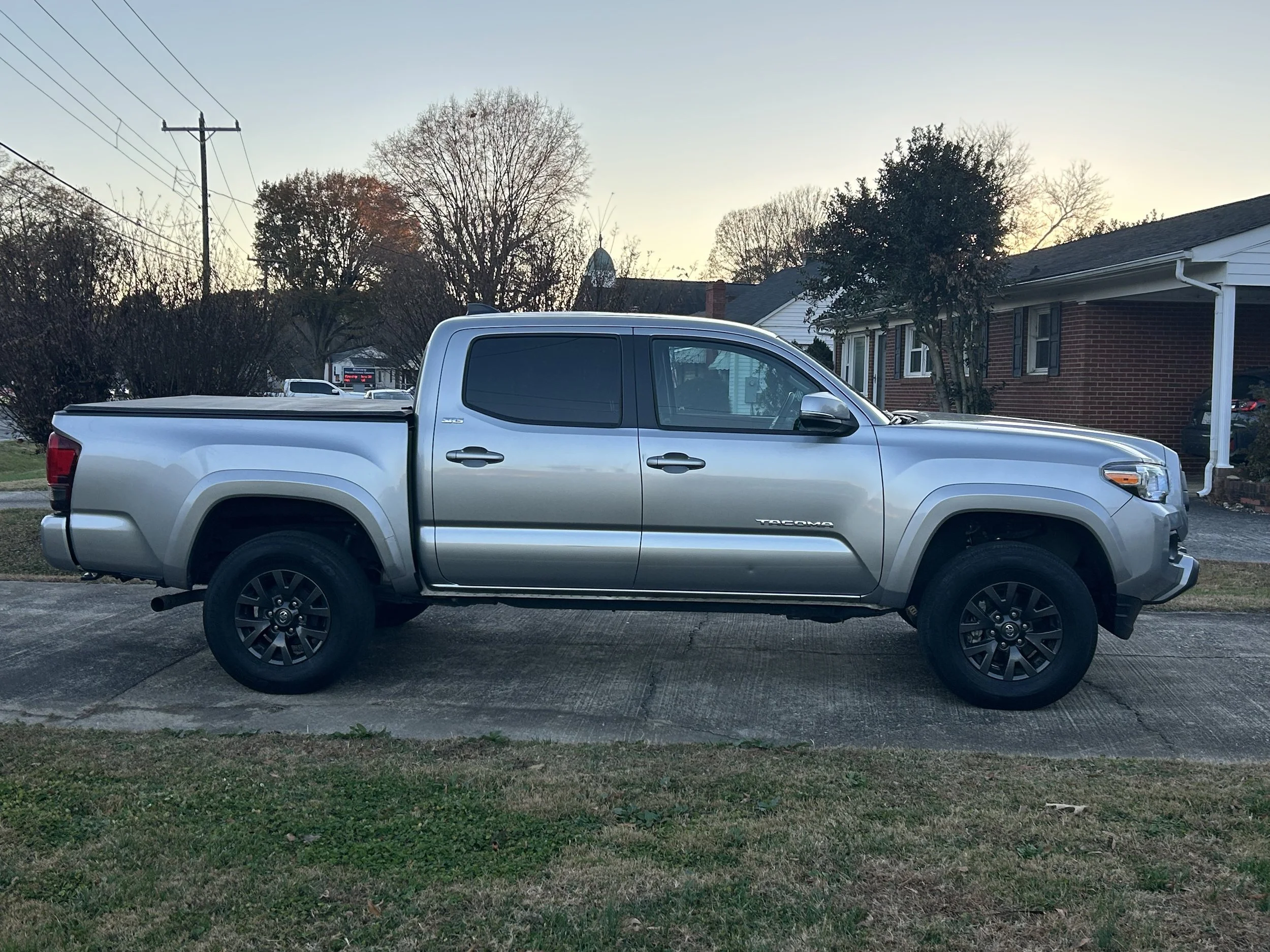 Side view of a silver Toyota Tacoma pickup truck parked on a driveway with houses and trees in the background.