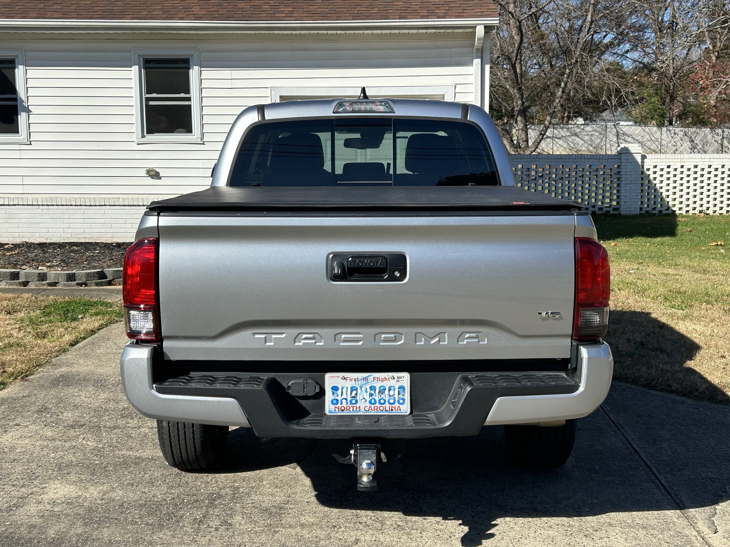Rear view of a silver Toyota Tacoma pickup truck parked on a concrete driveway in front of a white house with three windows, a flower bed, and a white lattice fence, with trees in the background.