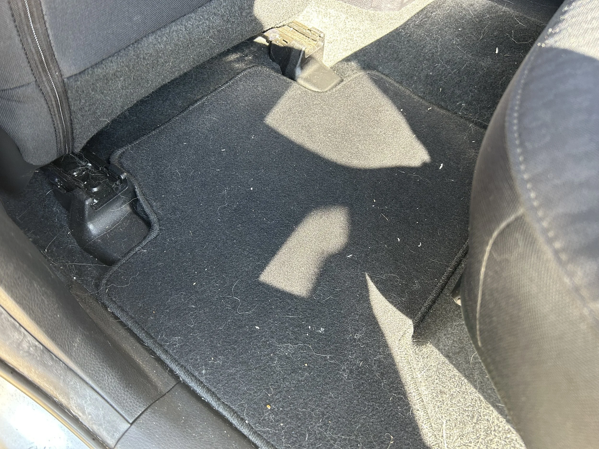 The back seat area of a car showing the floor mat, seat bolts, and a shadow cast on the mat.