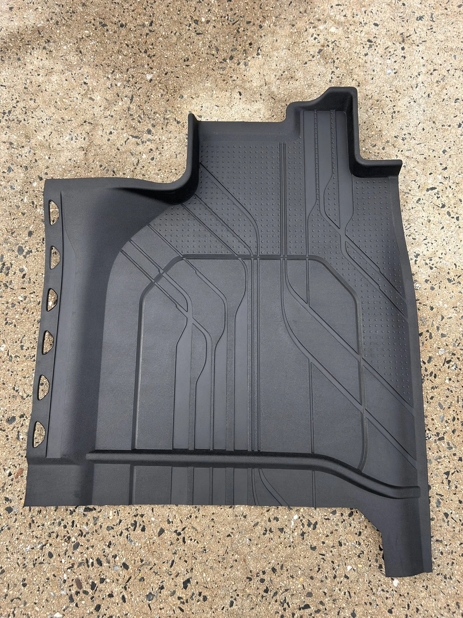 Black rubber car floor mat with a textured design on a speckled brown surface.
