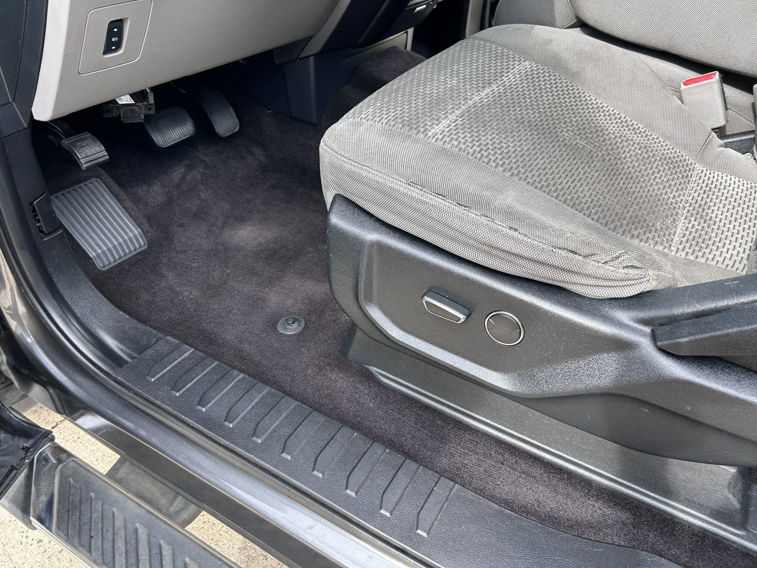 The image shows the interior driver side of a vehicle. Visible are the pedals, seat adjustment controls, the edge of the driver's seat, and the vehicle's carpeted floor.