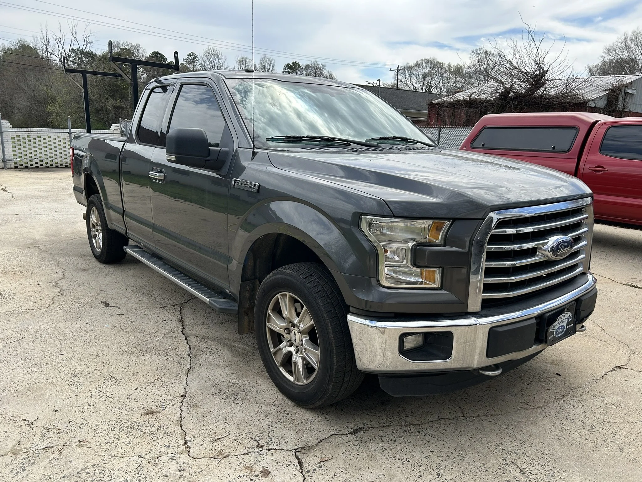 Gray Ford F-150 pickup truck parked on concrete surface next to a red truck, with a chain-linked fence and trees in the background.