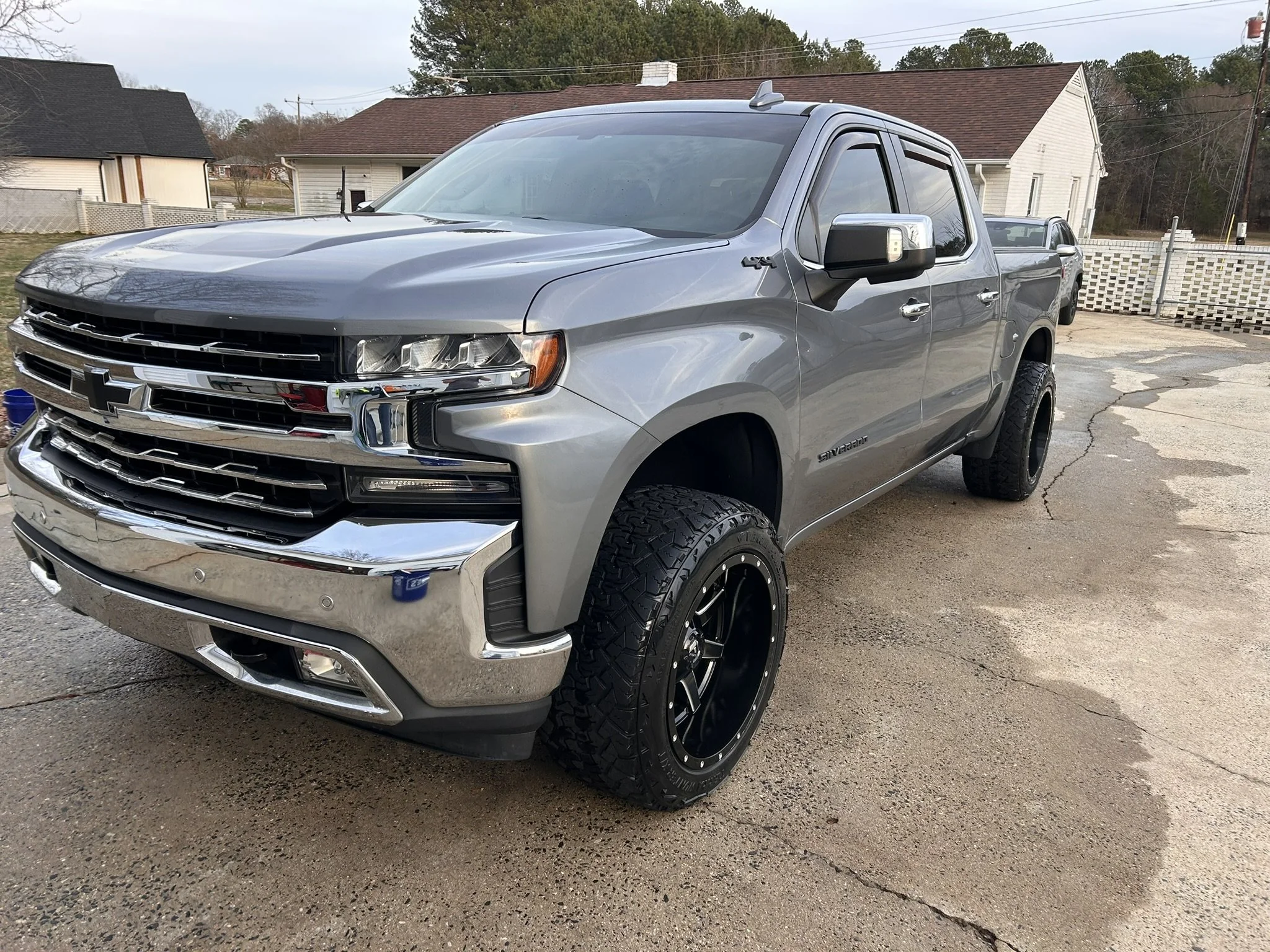 Silver Chevrolet Silverado pickup truck parked on a driveway with house and trees in the background.