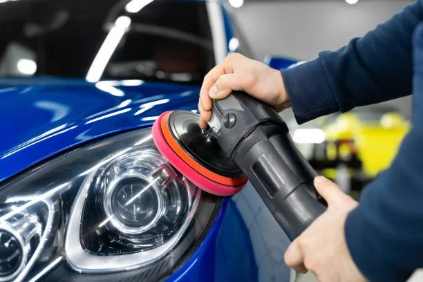 Person polishing a blue car headlight with a handheld electric buffer.