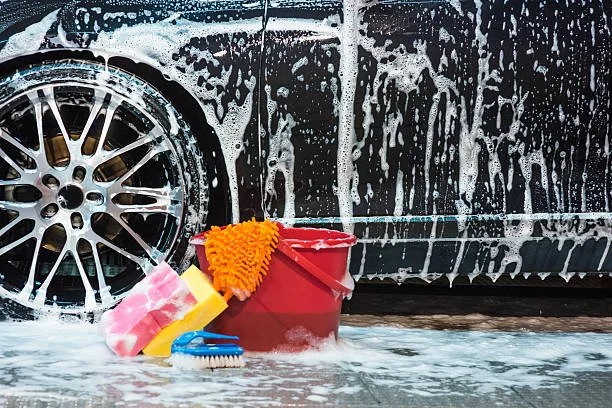 Car being washed with a soap-soaked sponge, brush, and bucket of water in front of the car's side with soap suds and foam.