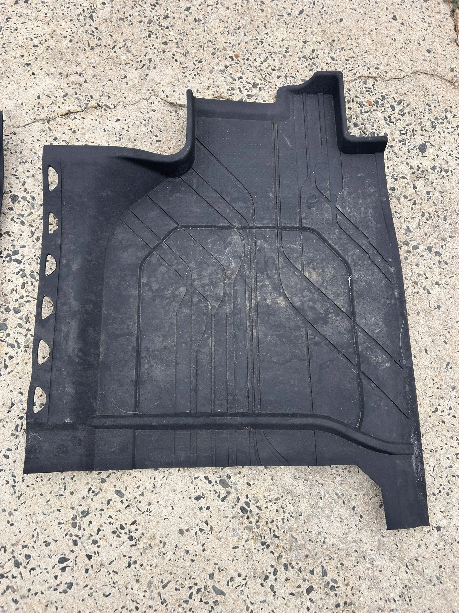 A black rubber car floor mat with grooves, placed on a concrete surface.
