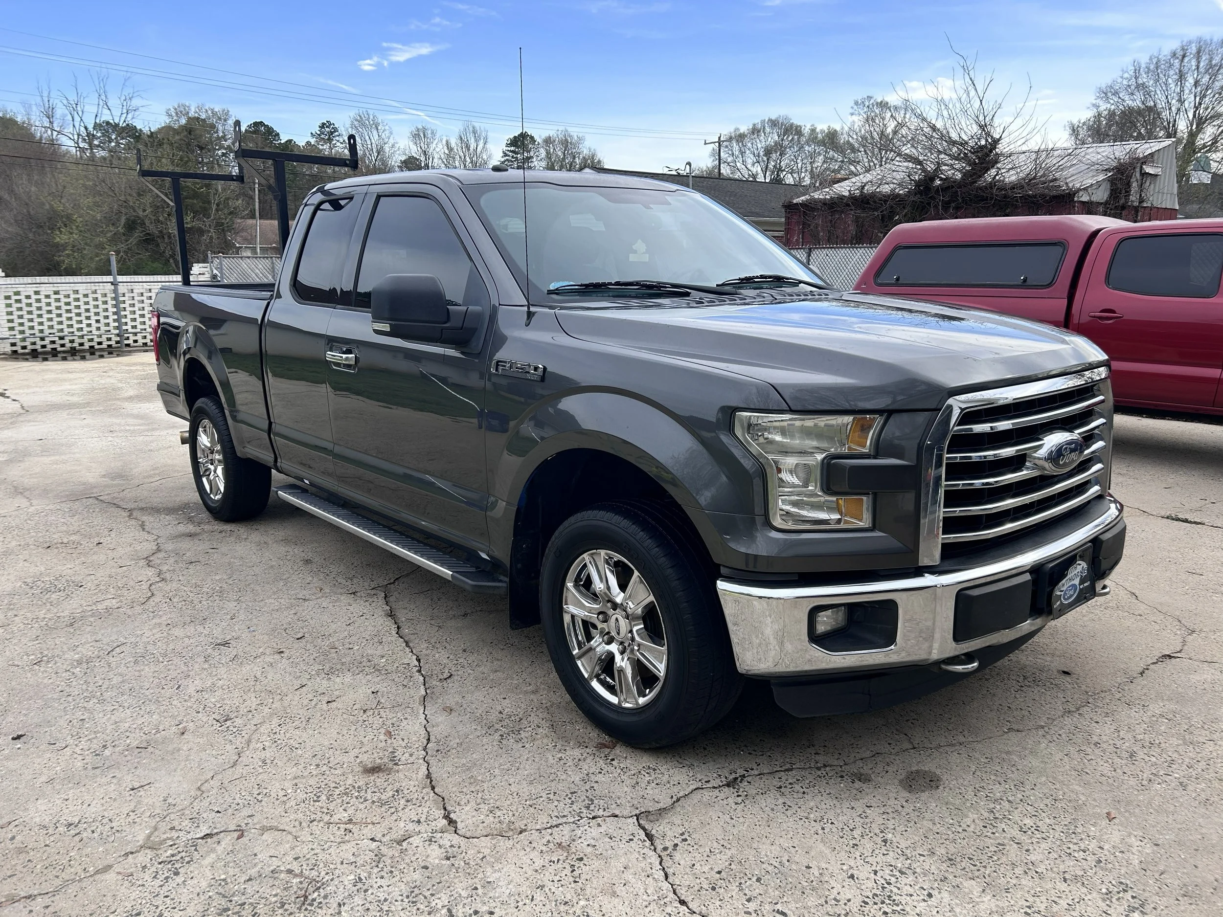 Gray Ford F-150 pickup truck parked on cracked asphalt, with a red truck and a barn in the background, under partly cloudy sky.
