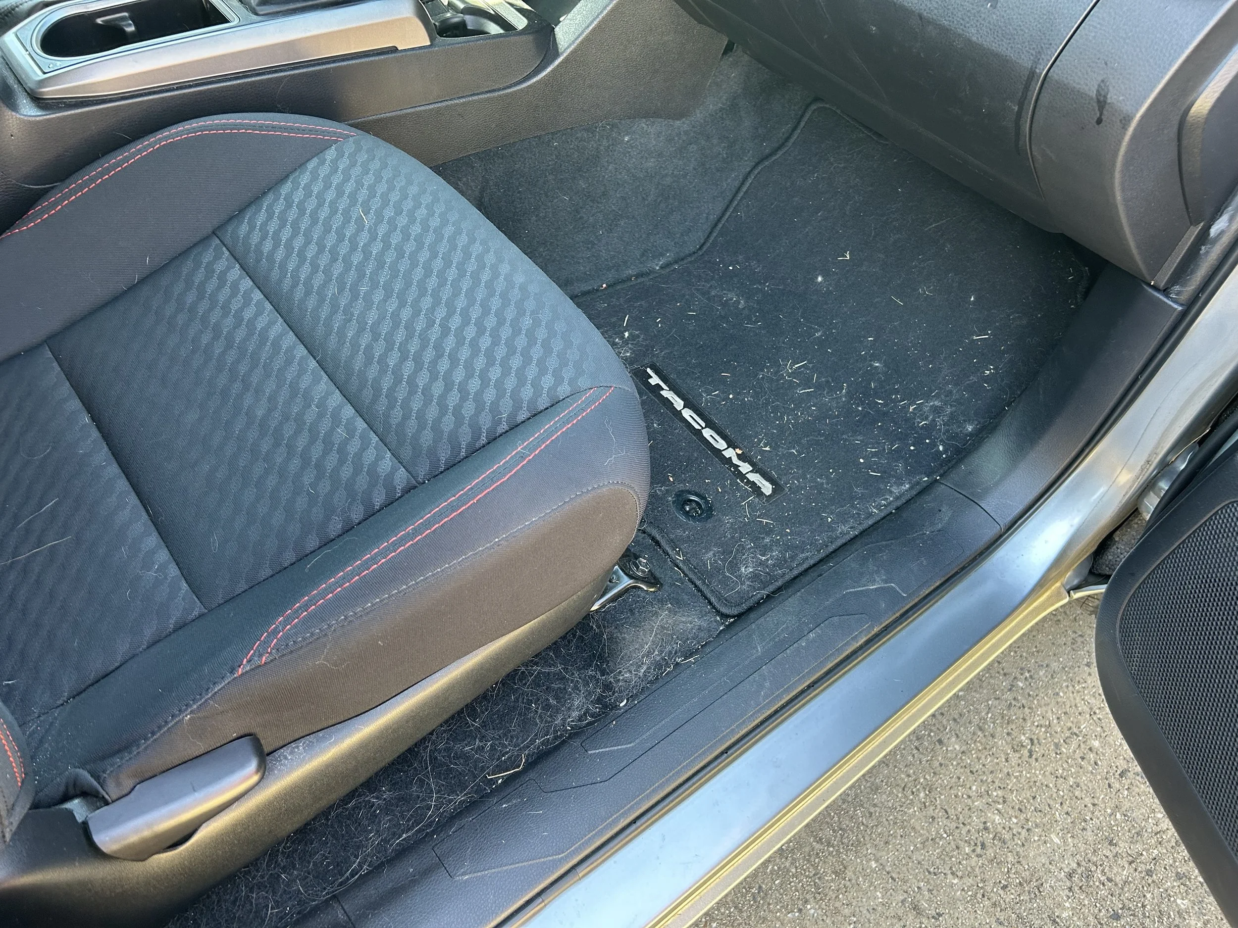 Inside view of a car showing a black fabric passenger seat with red stitching, a black floor mat with a 
