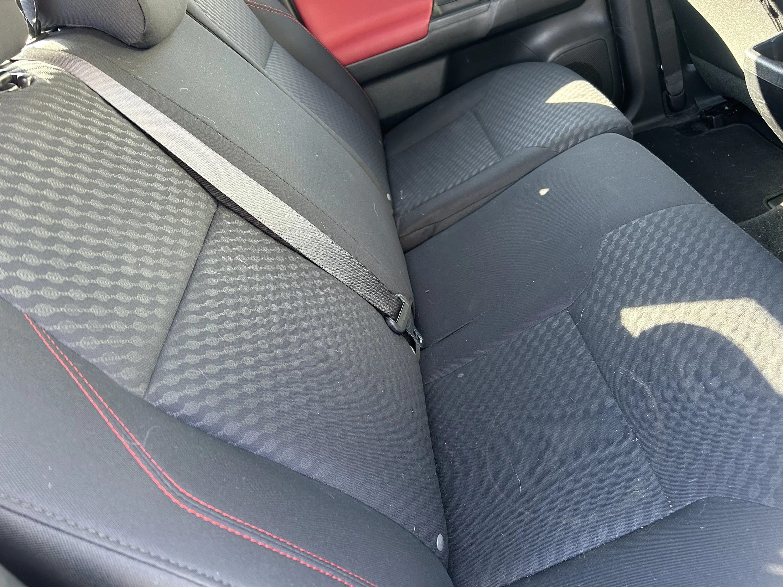 The backseat of a car showing black fabric seats with a textured pattern, a seatbelt, and some small dirt and hair particles.