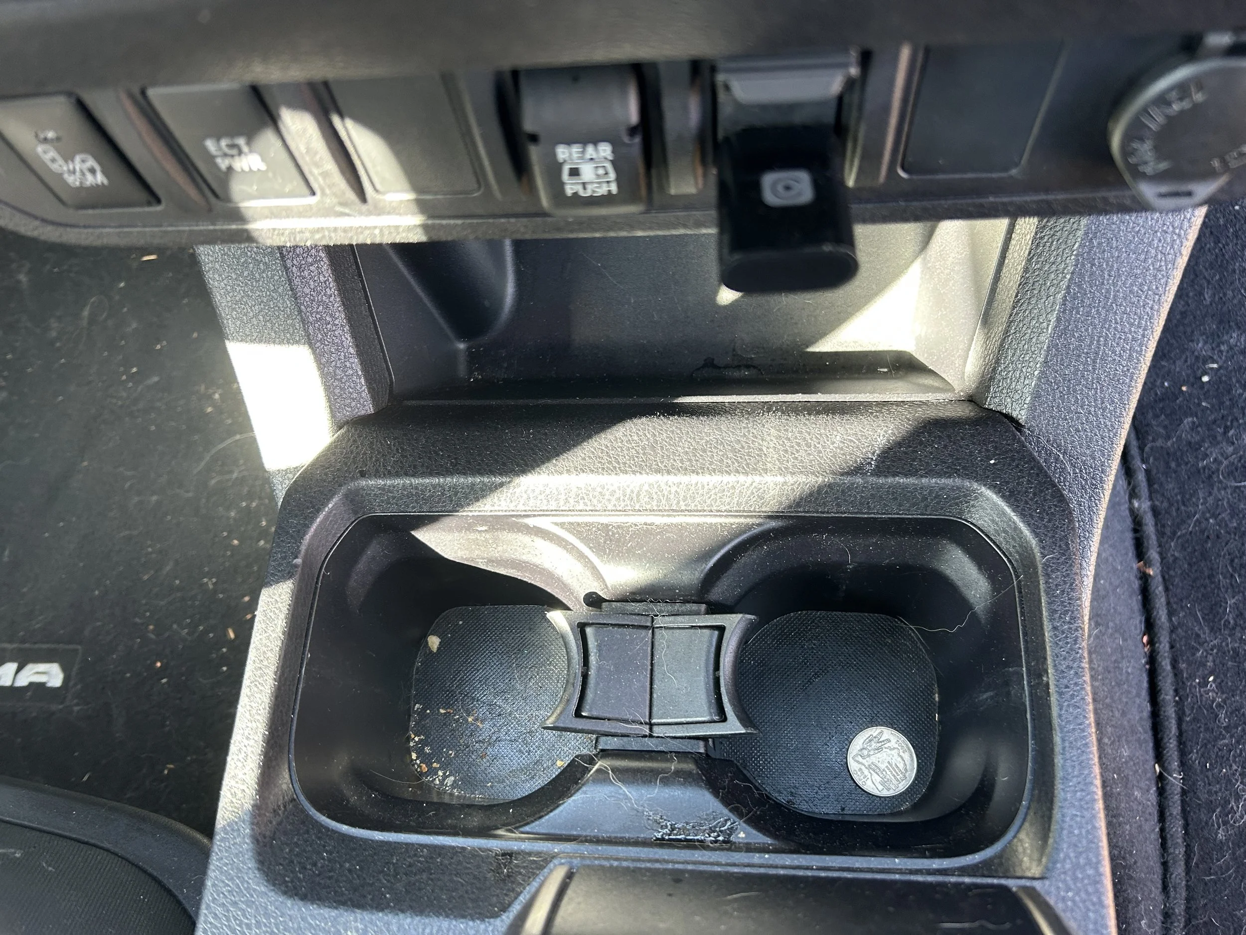 Car cup holders with dirt and crumbs inside. There's a small round item, possibly a coin or token, in the right cup holder.