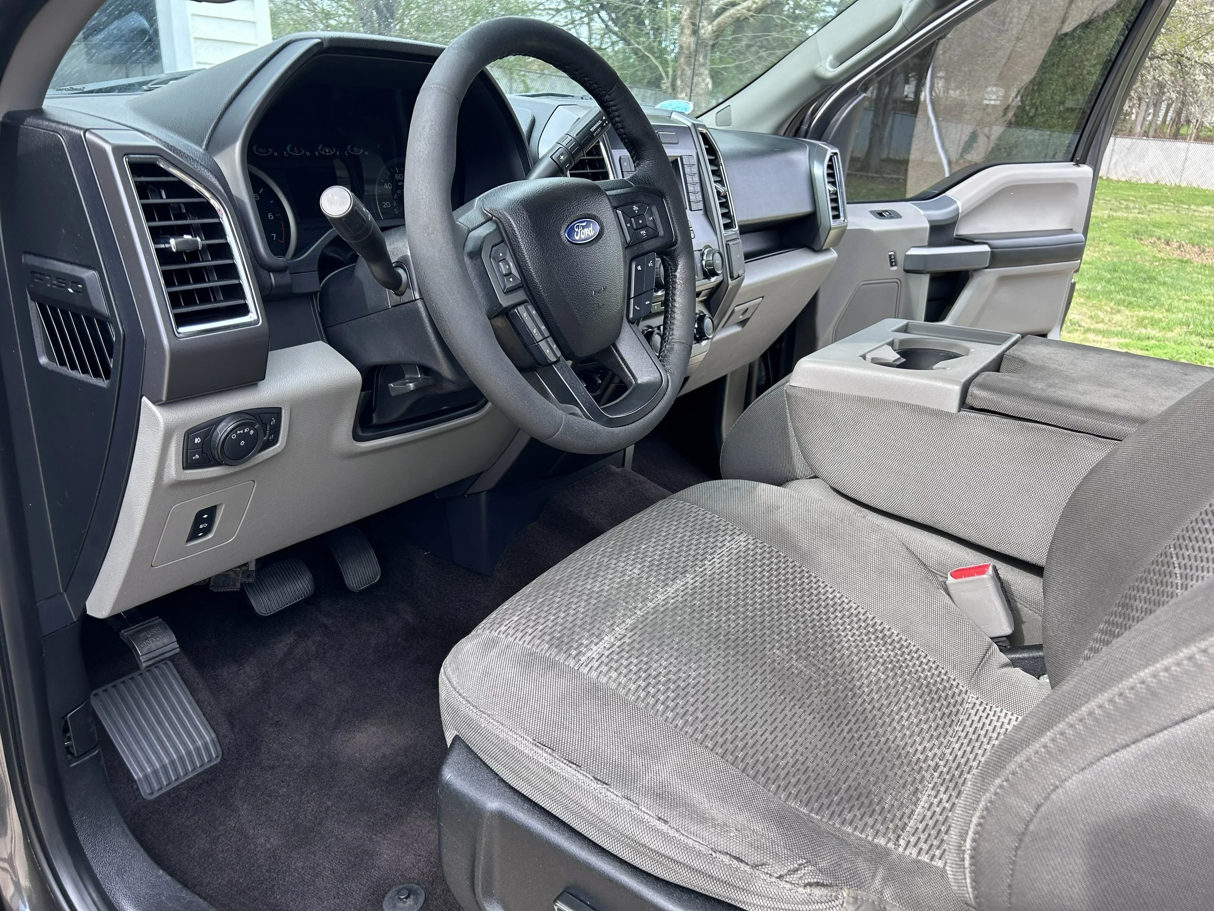 Interior view of a Ford truck's driver's side, showing the steering wheel, dashboard, pedals, gray fabric seat, center console, and passenger side door with a window, set against a background of green grass and trees.