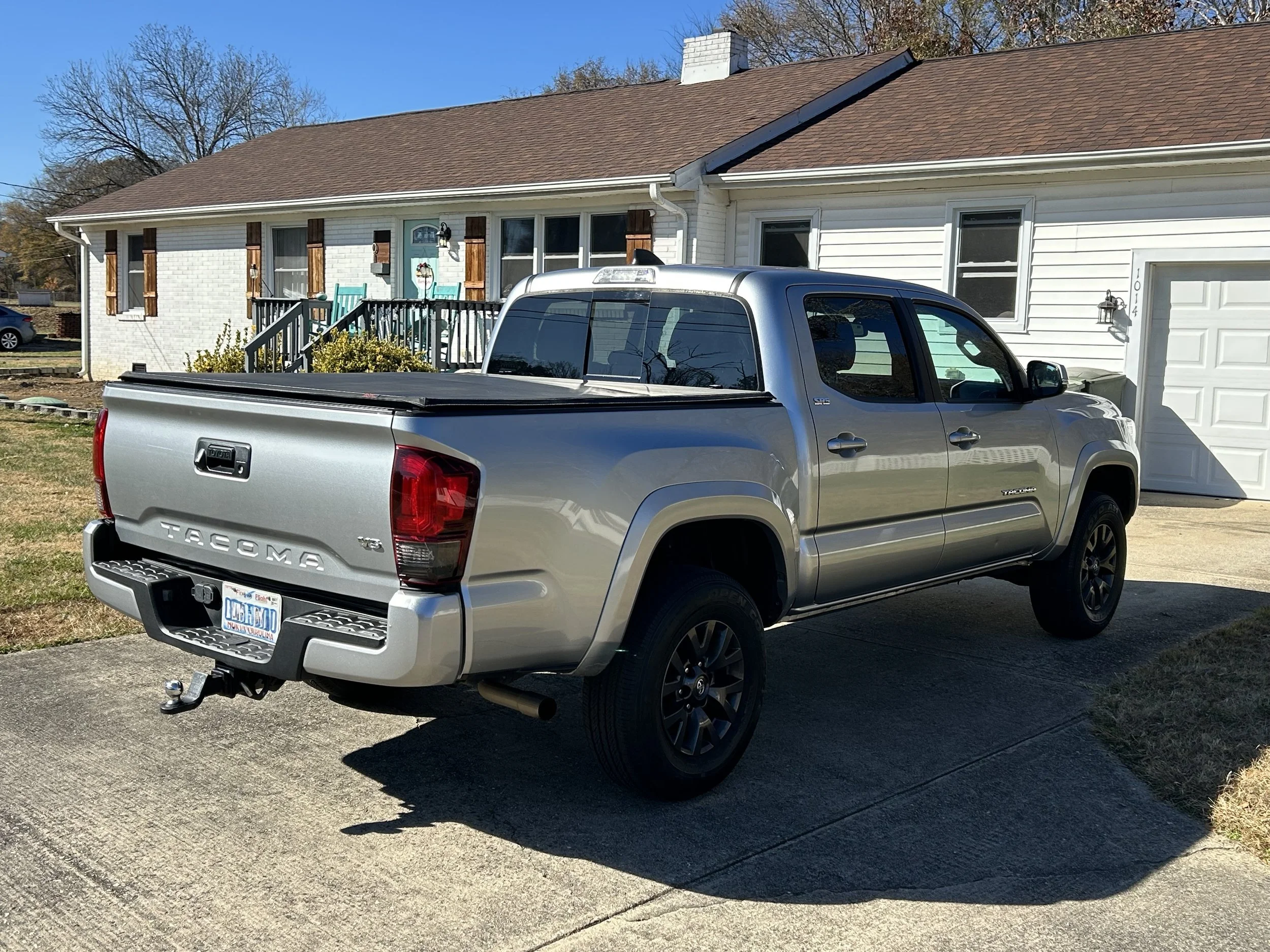 Silver Toyota Tacoma pickup truck parked in driveway in front of a white house with brown shutters.