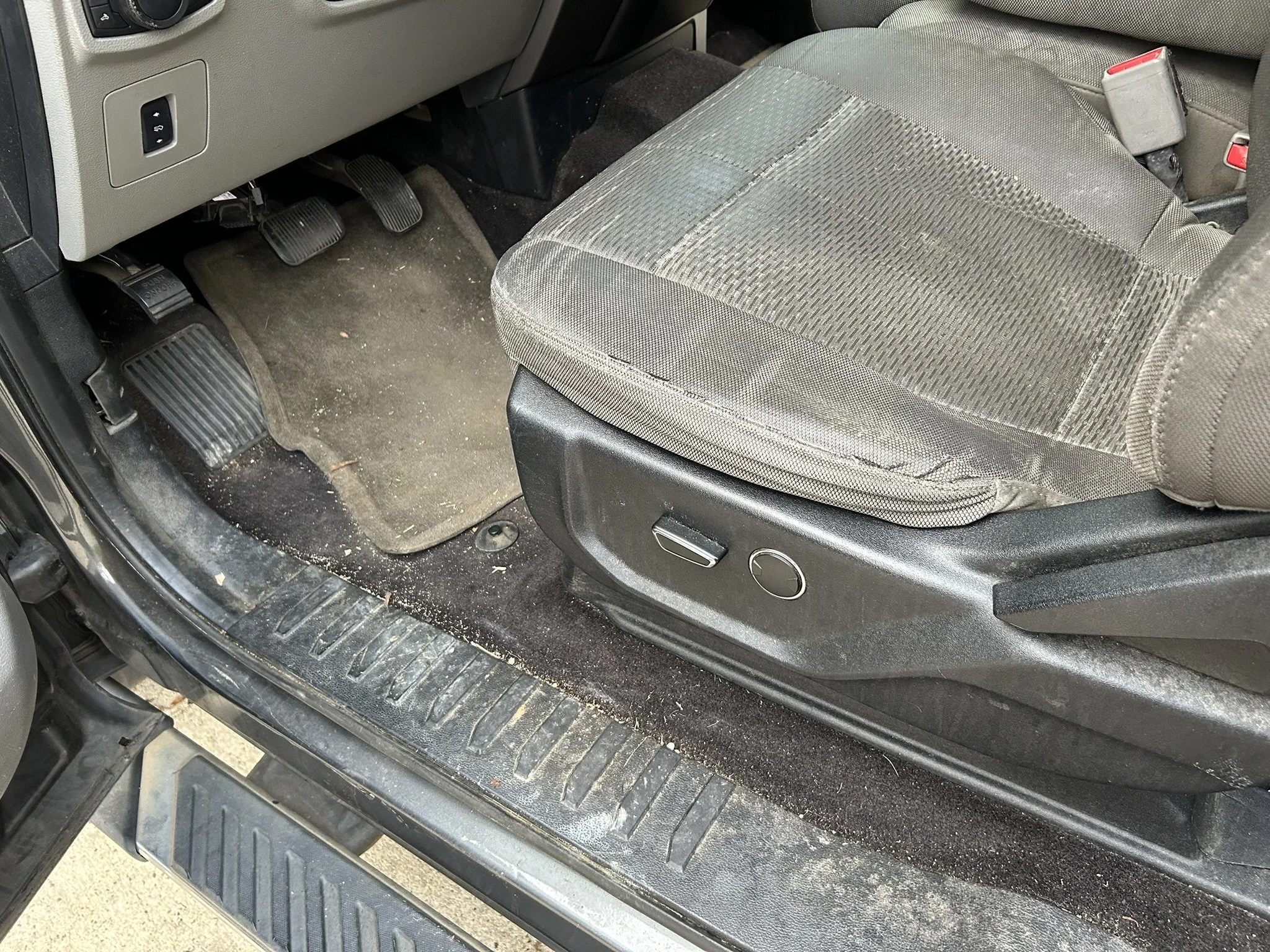 The interior of a vehicle showing the driver's side floor area with a dirty carpet, two pedals, and part of the seat with side controls.