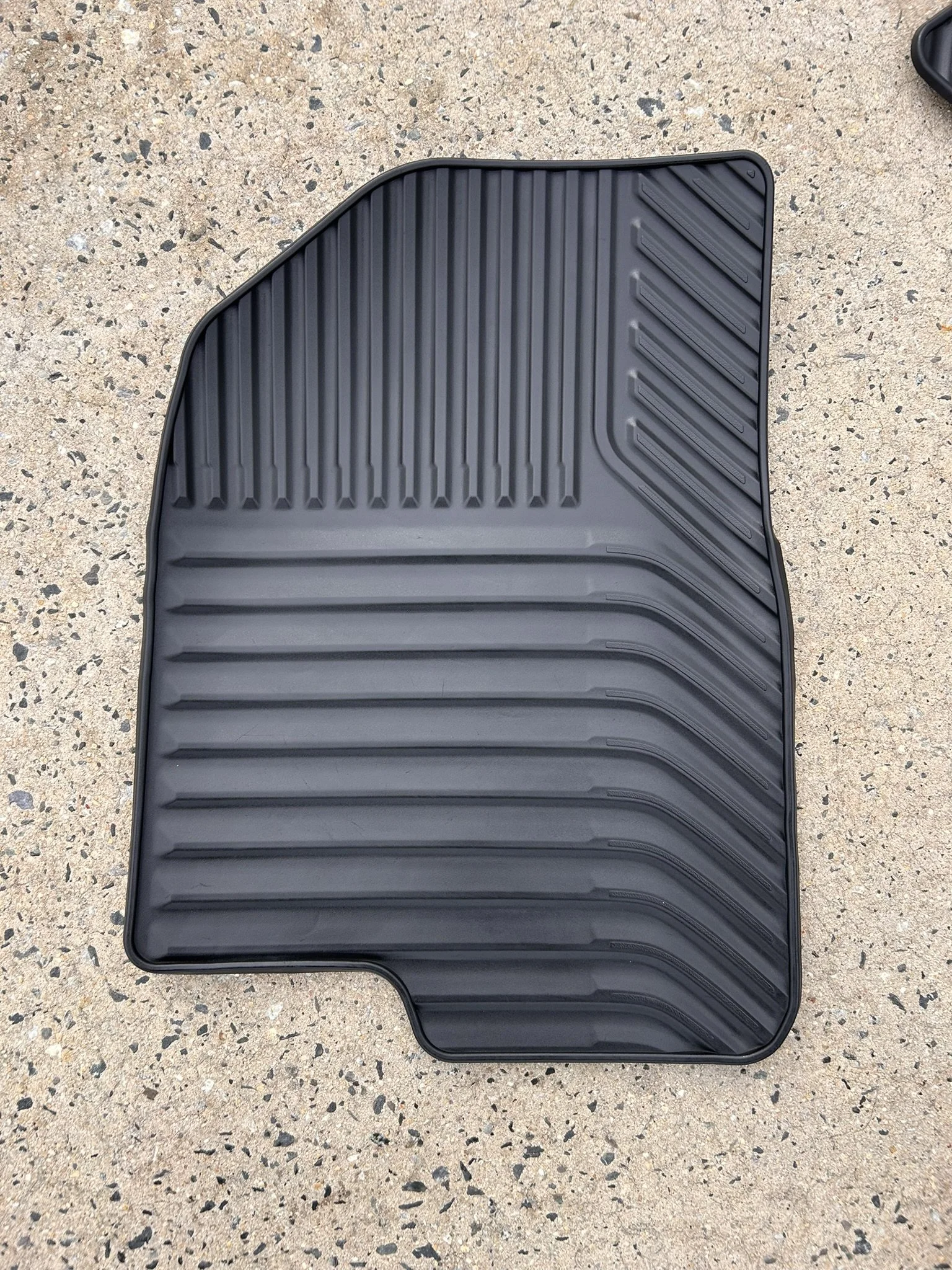 Black rubber car floor mat with ridged design on a speckled concrete surface.
