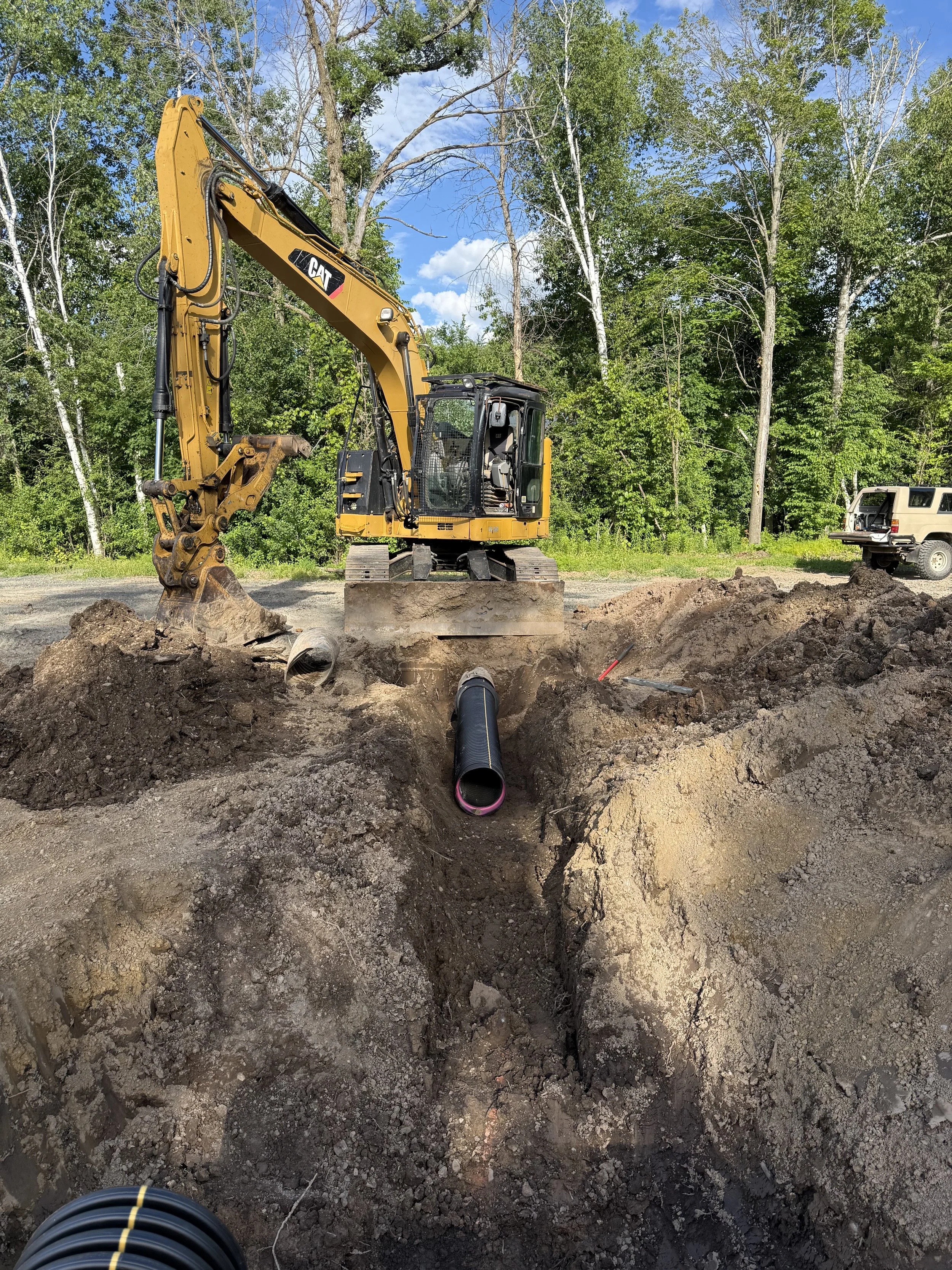 Construction site with a yellow excavator digging a trench for underground piping, involving black pipes.