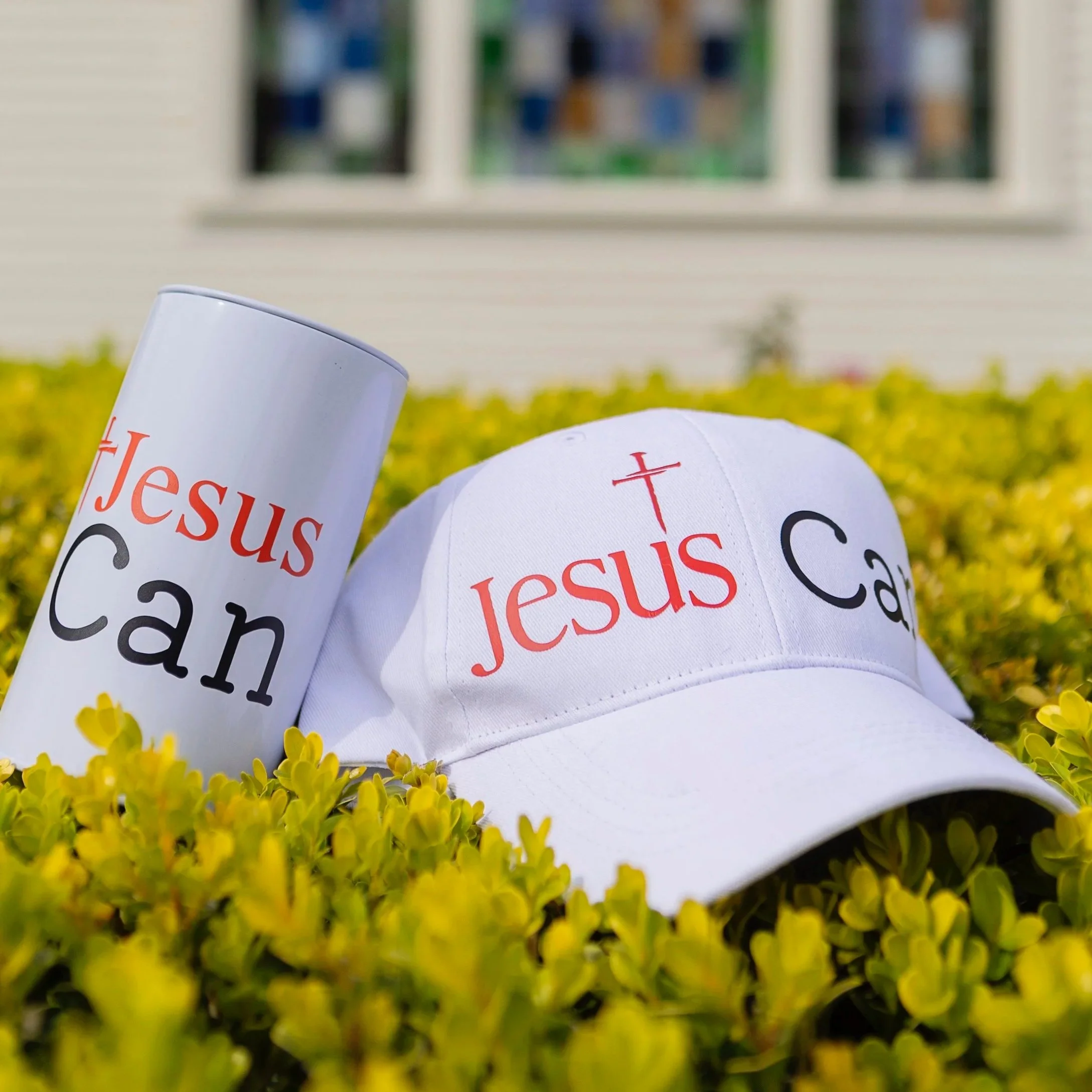 A white cap and a white tumbler with the words 'Jesus Can' in red and black, placed among yellow bushes outside a house with white siding and a window.