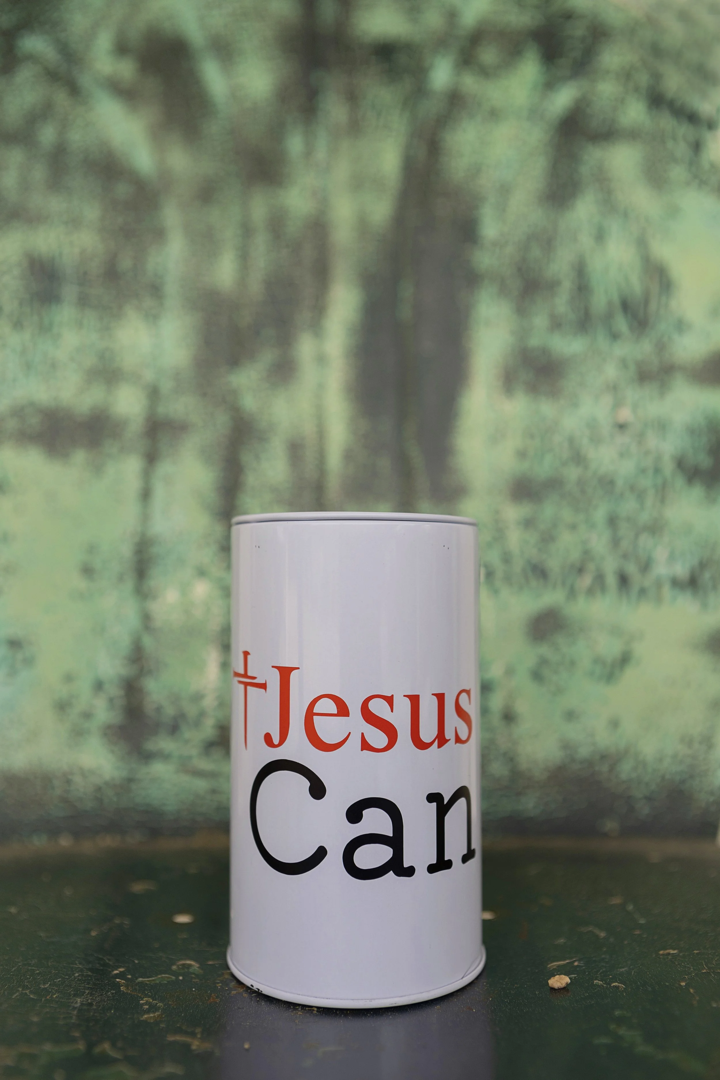 A white can with the text 'Jesus Can' printed on it, placed on a dark surface with a blurred green forest background.