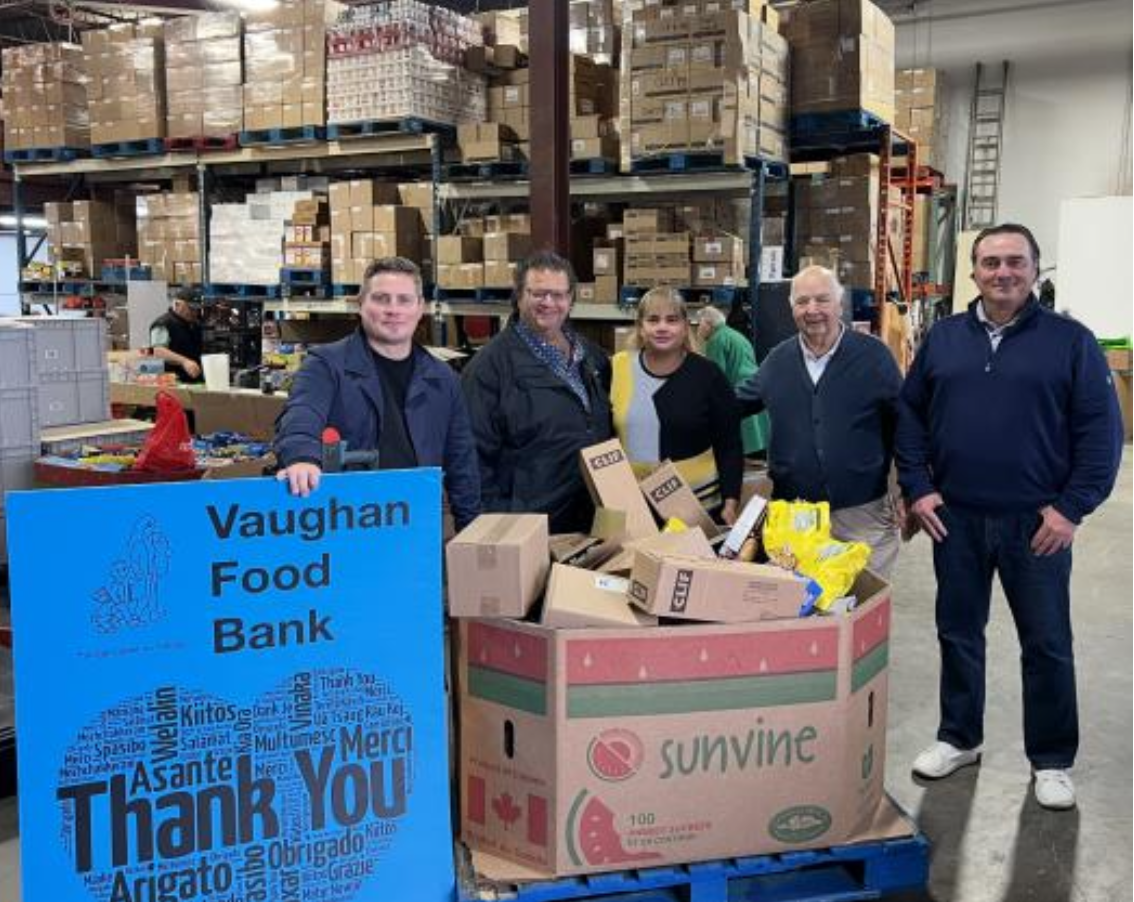 a group of people smiling surrounded by boxes and holding a sign for the Vaughan Food Bank