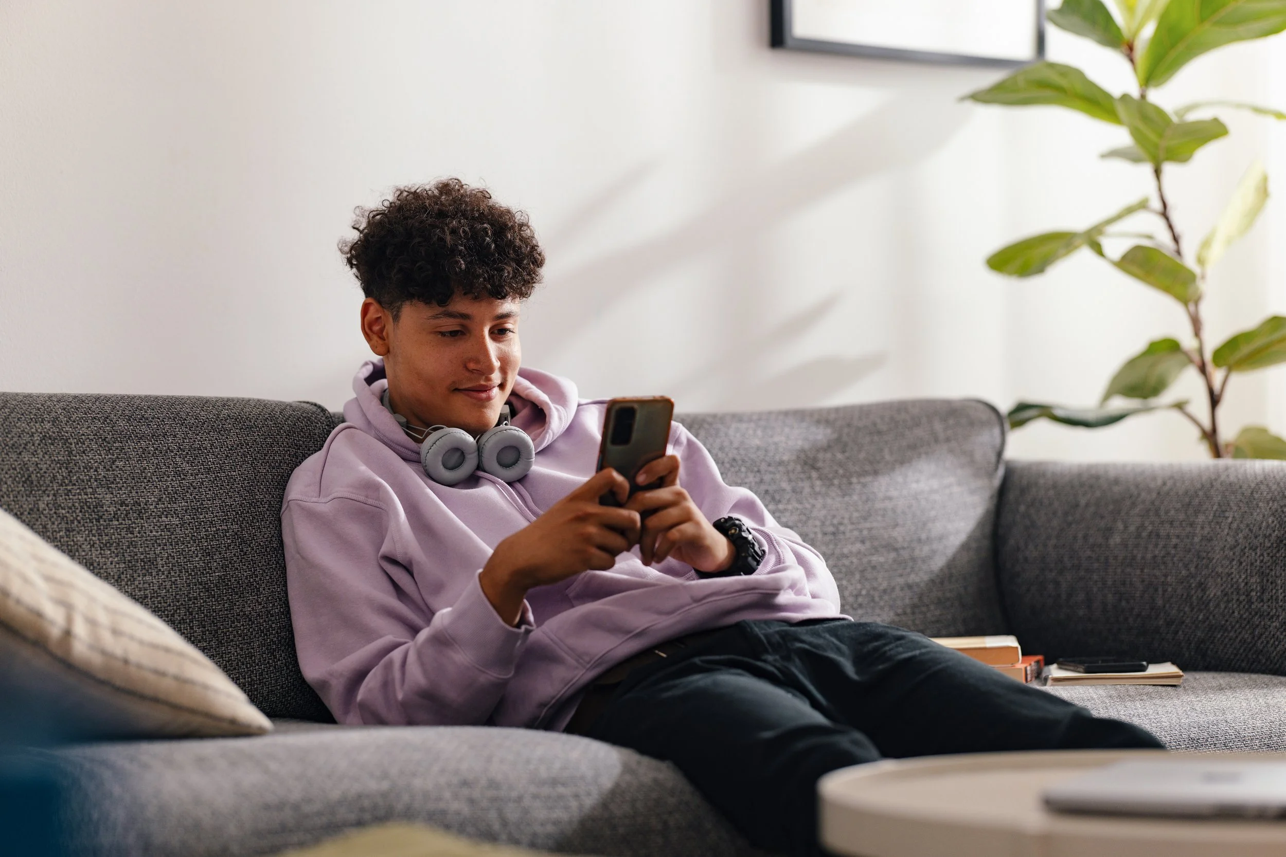 A young man with curly hair wearing a lavender hoodie and gray headphones around his neck is sitting on a gray sofa, looking at his smartphone, in a bright living room with a large plant and a framed picture on the wall.
