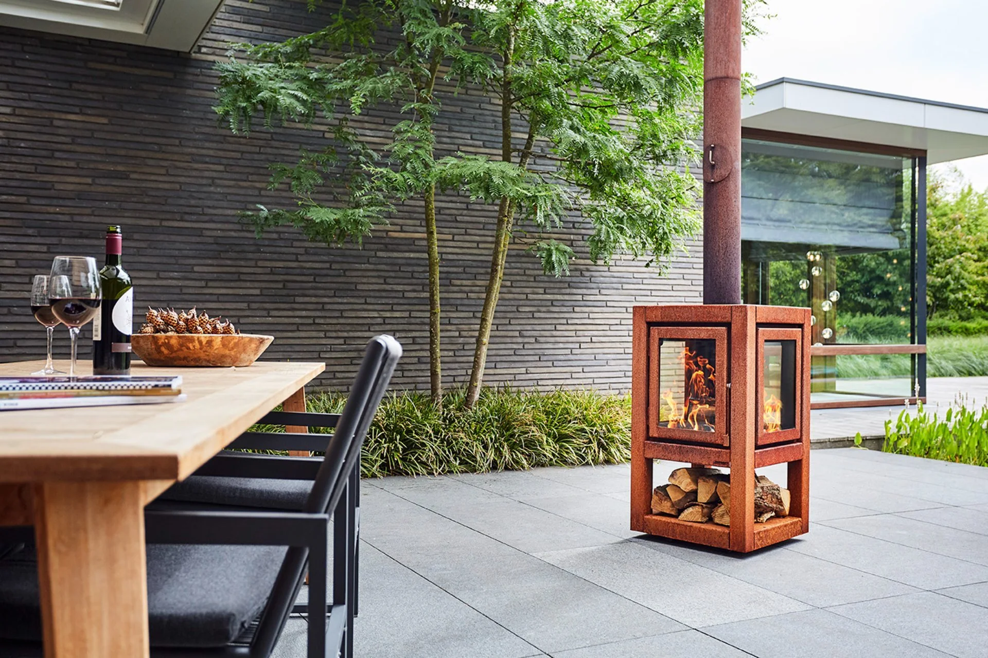 Outdoor Terrasse mit einem Feuerkorb, Holz und einem Tisch mit Rotwein, Weingläsern und einer Schale mit Walnüssen vor einem modernen Haus mit brauner Ziegelmauer und großen Fenstern.