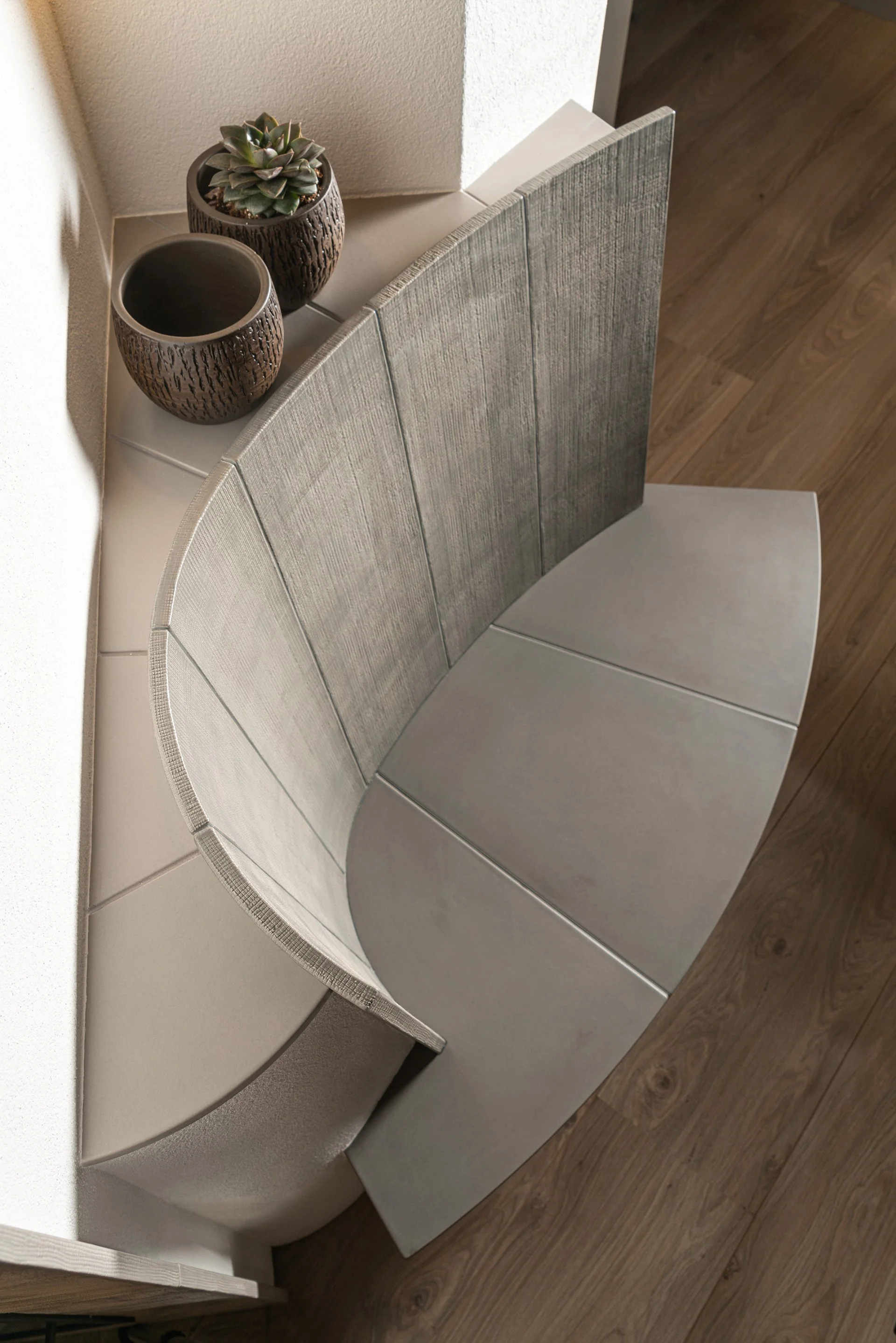 Ein halbkreisförmiger Hocker mit silberner Oberfläche und einem halbkreisförmigen Rückenlehnen-Design, daneben zwei braune Keramiktöpfe mit Sukkulenten, auf einer weißen Tischplatte in einem Raum mit Holzboden.