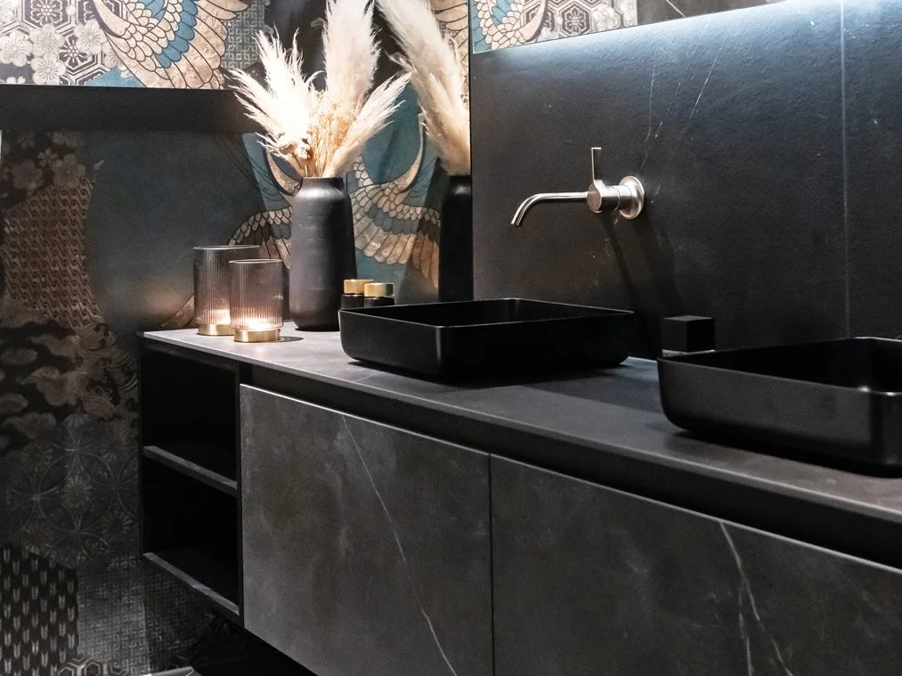 Ein schwarzer Waschtisch mit einem Wasserhahn, zwei schwarze Behälter und Kerzen auf der Oberfläche, im Hintergrund eine Wand mit metallicfarbenen, dekorativen Mustern und eine Vase mit getrockneten Pflanzen.