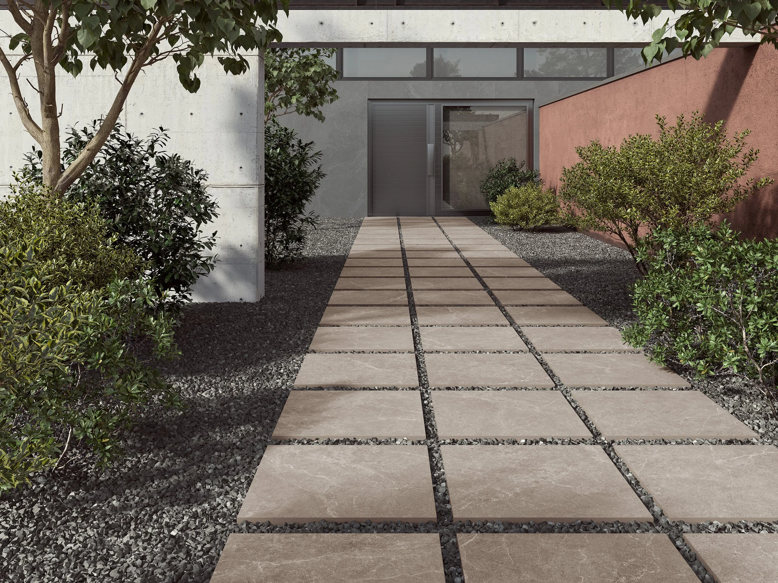 Moderne Gehweg aus quadratischen Steinen mit dunklen Fugen, umgeben von kleinen dunklen Kieselsteinen, vor einem modernen Haus mit grauer und roter Wand, sowie vielen grünen Sträuchern und Bäumen.