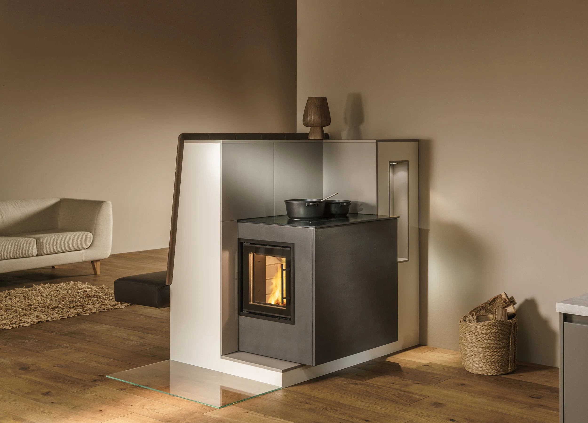 Moderne Feuerstelle in einer Wohnküche, daneben ein Korb mit Holzscheiten, im Hintergrund ein Sofa und ein Teppich.