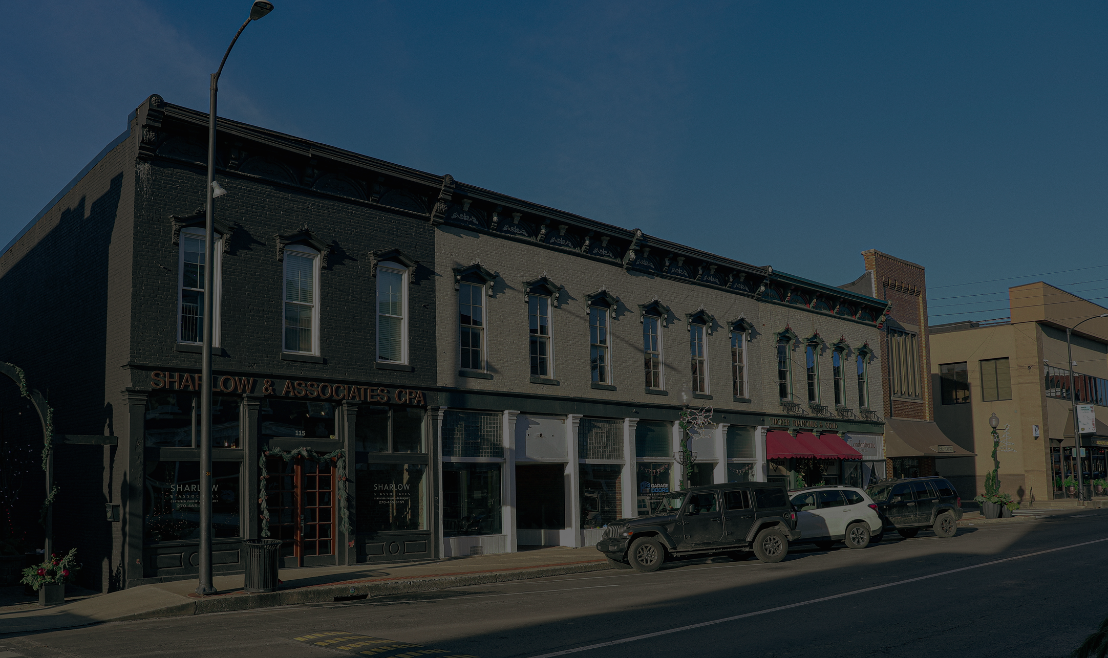 Photograph of a commercial building with multiple business storefronts on a city street during daytime, including a CPA office and a jewelry store, with parked cars and a streetlamp in the foreground.