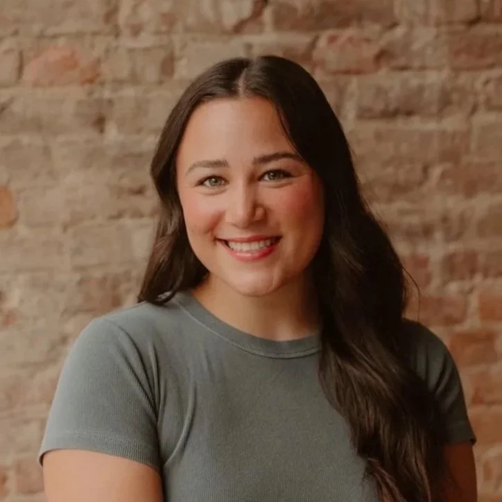 A young woman with long dark hair and a bright smile, wearing a gray t-shirt, standing in front of a brick wall.